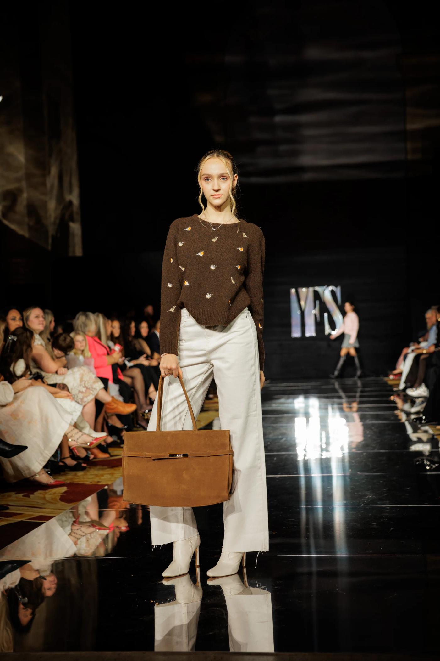 A model in a brown knit and white trousers walking the YFS runway with a leather bag
