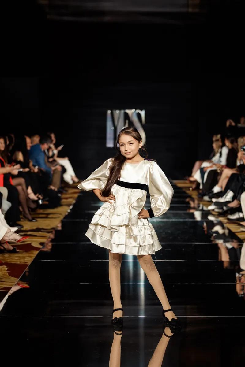 A girl in an ivory satin Petite Maison Kids dress with a black bow on the YFS runway
