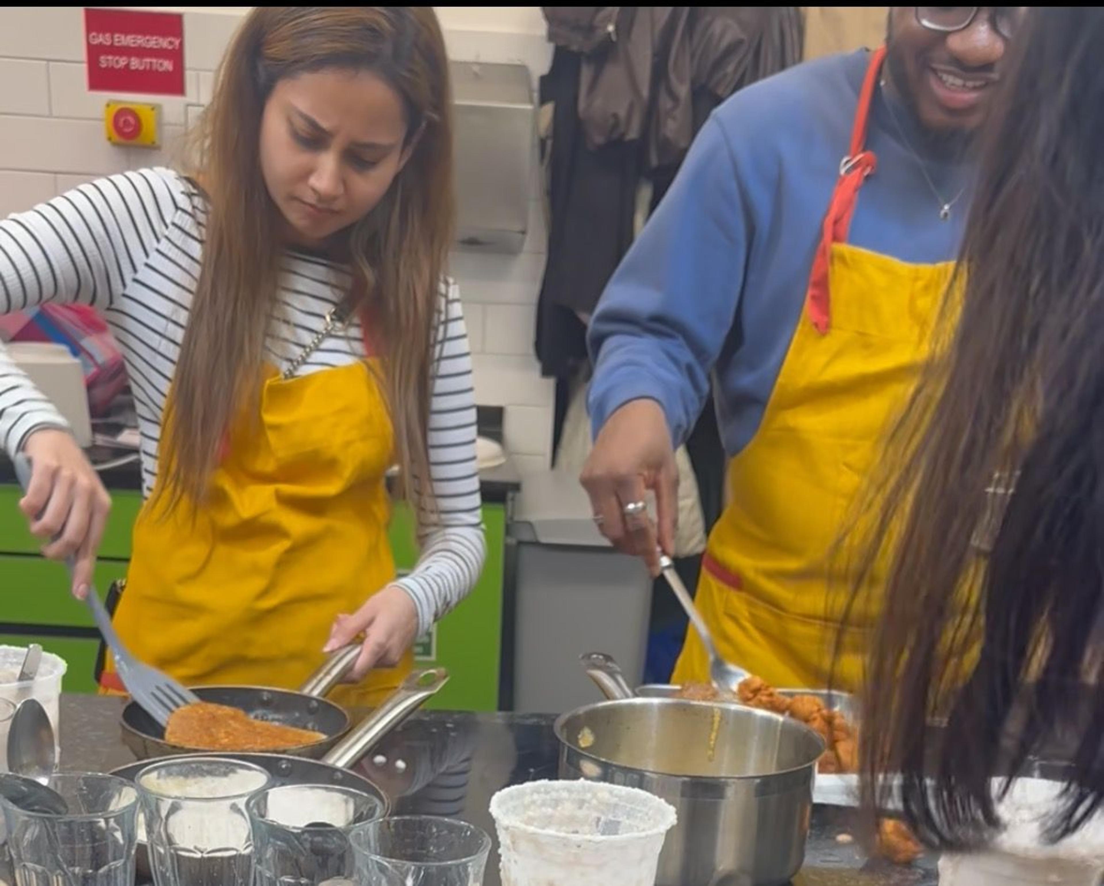 Two aproned guests frying patties and stirring a pot at a cooking-class station