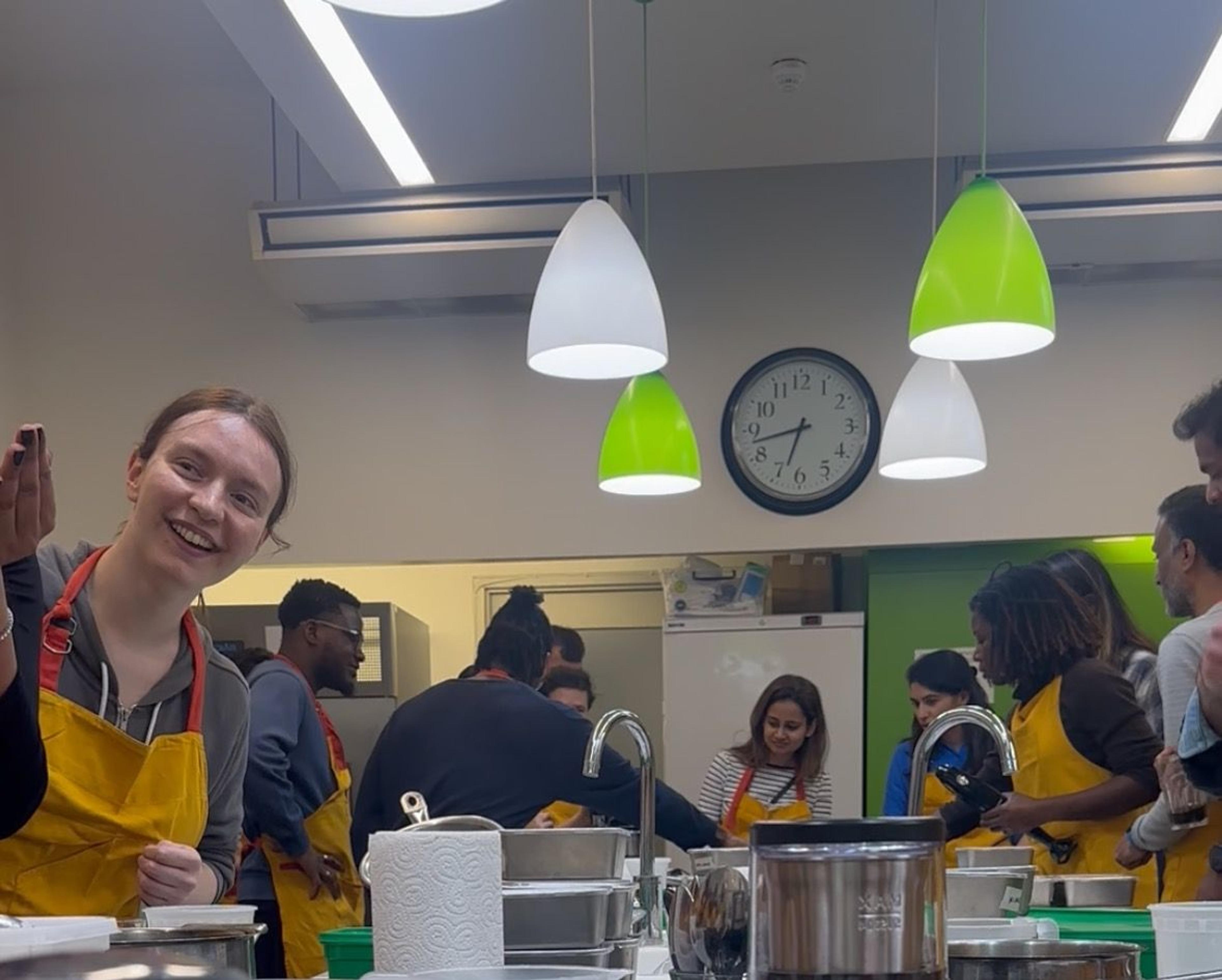 Smiling guest in a yellow apron at a busy cookery class, classmates cooking behind her