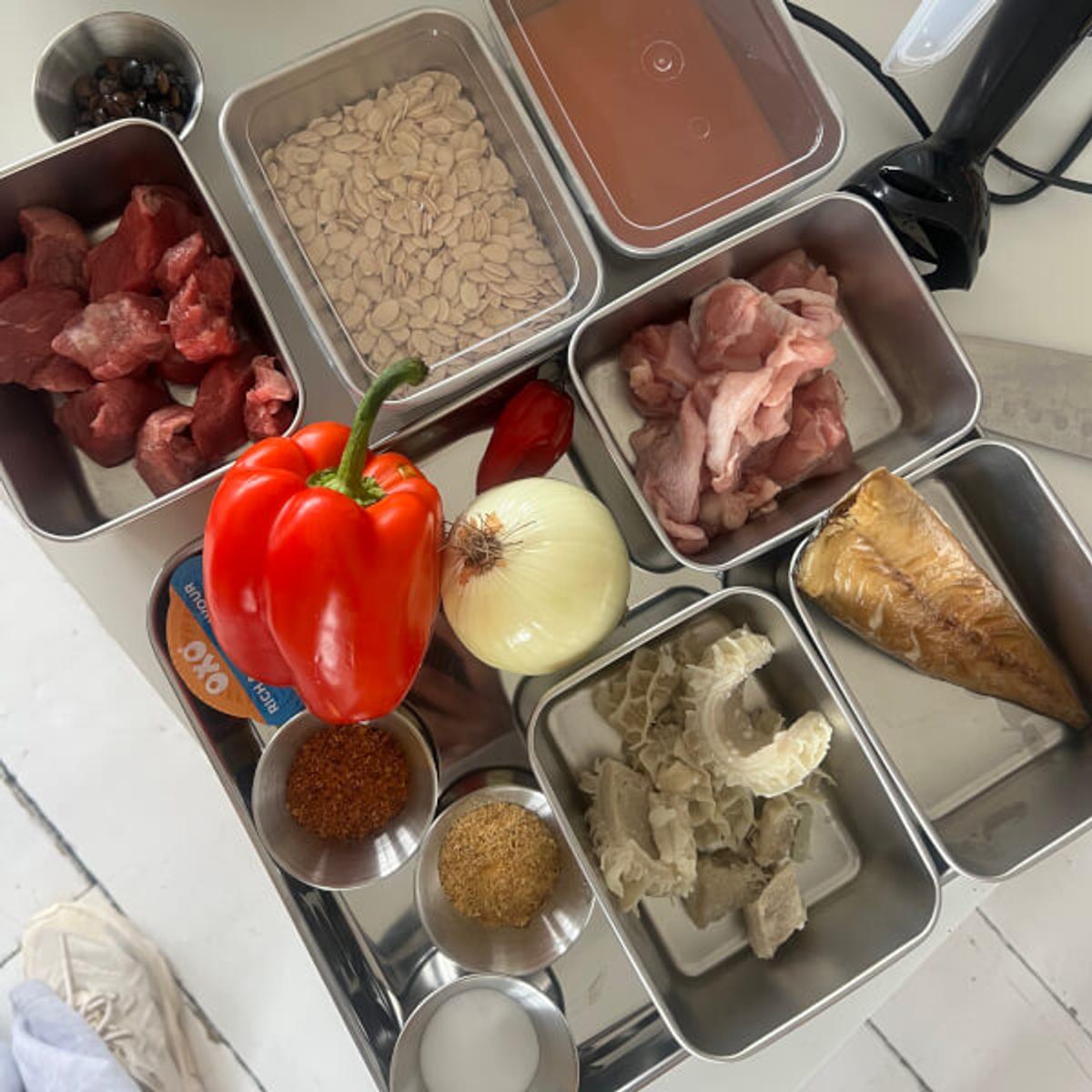 Flat-lay of West African cooking ingredients in metal trays: beef, egusi seeds, chicken, peppers and smoked fish