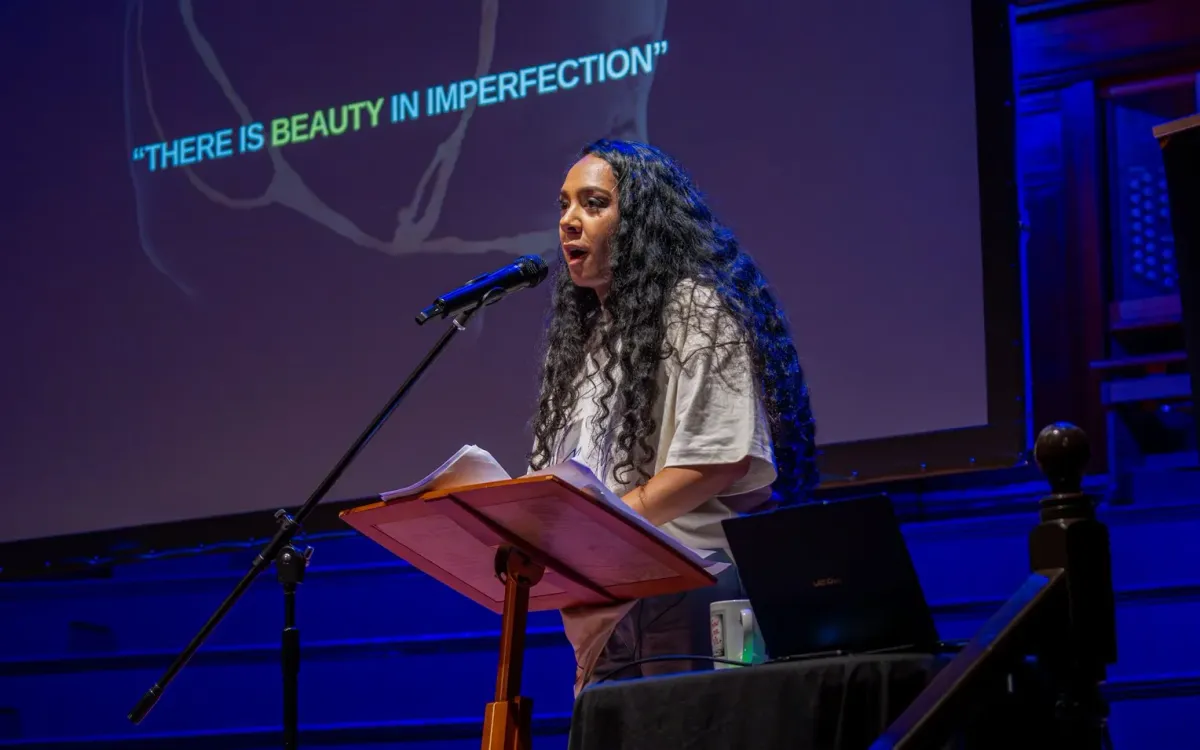 A woman speaking at a lectern on a stage lit blue, under the words There is beauty in imperfection