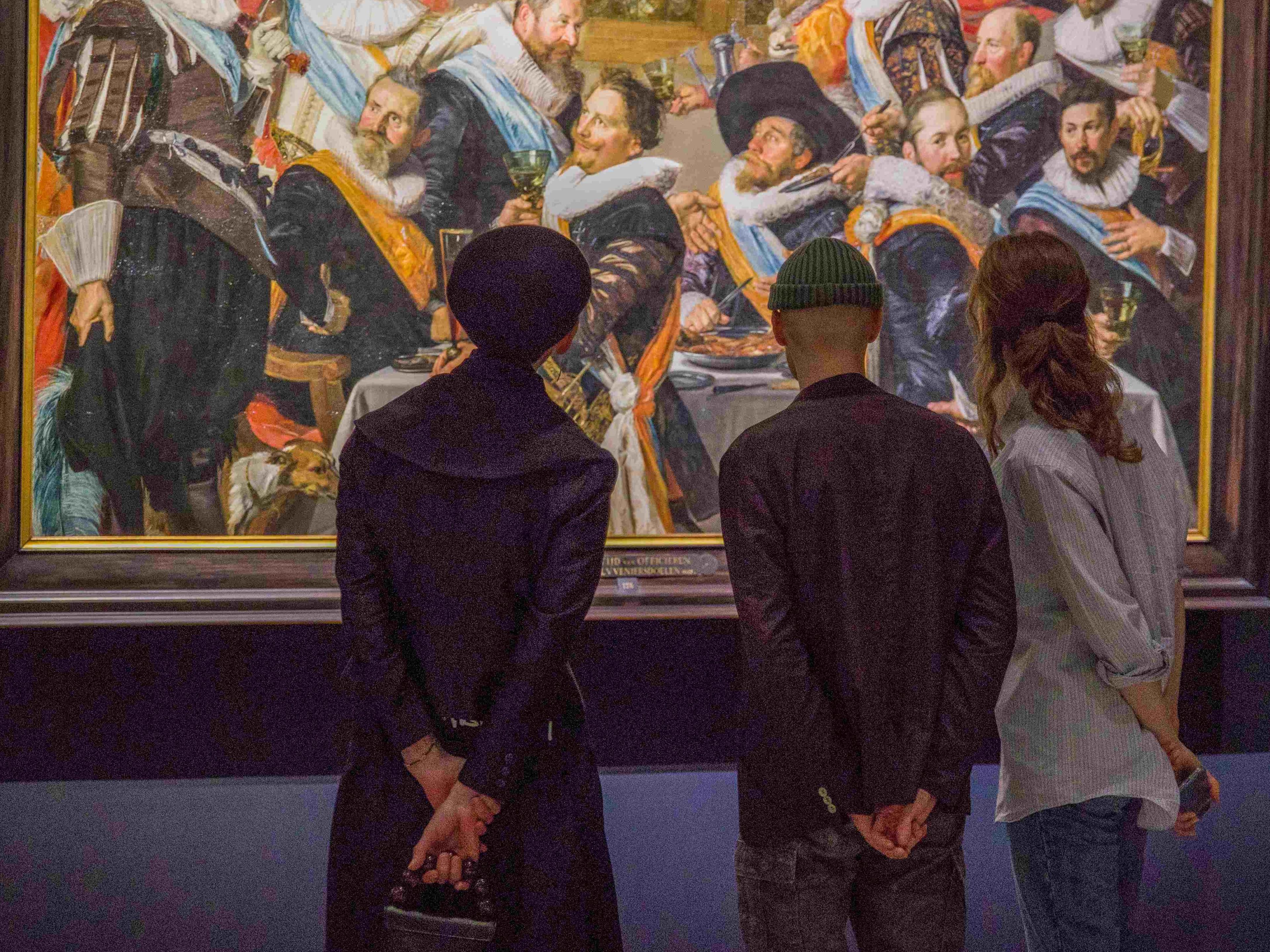 Drie bezoekers bekijken een schuttersstuk van Frans Hals.