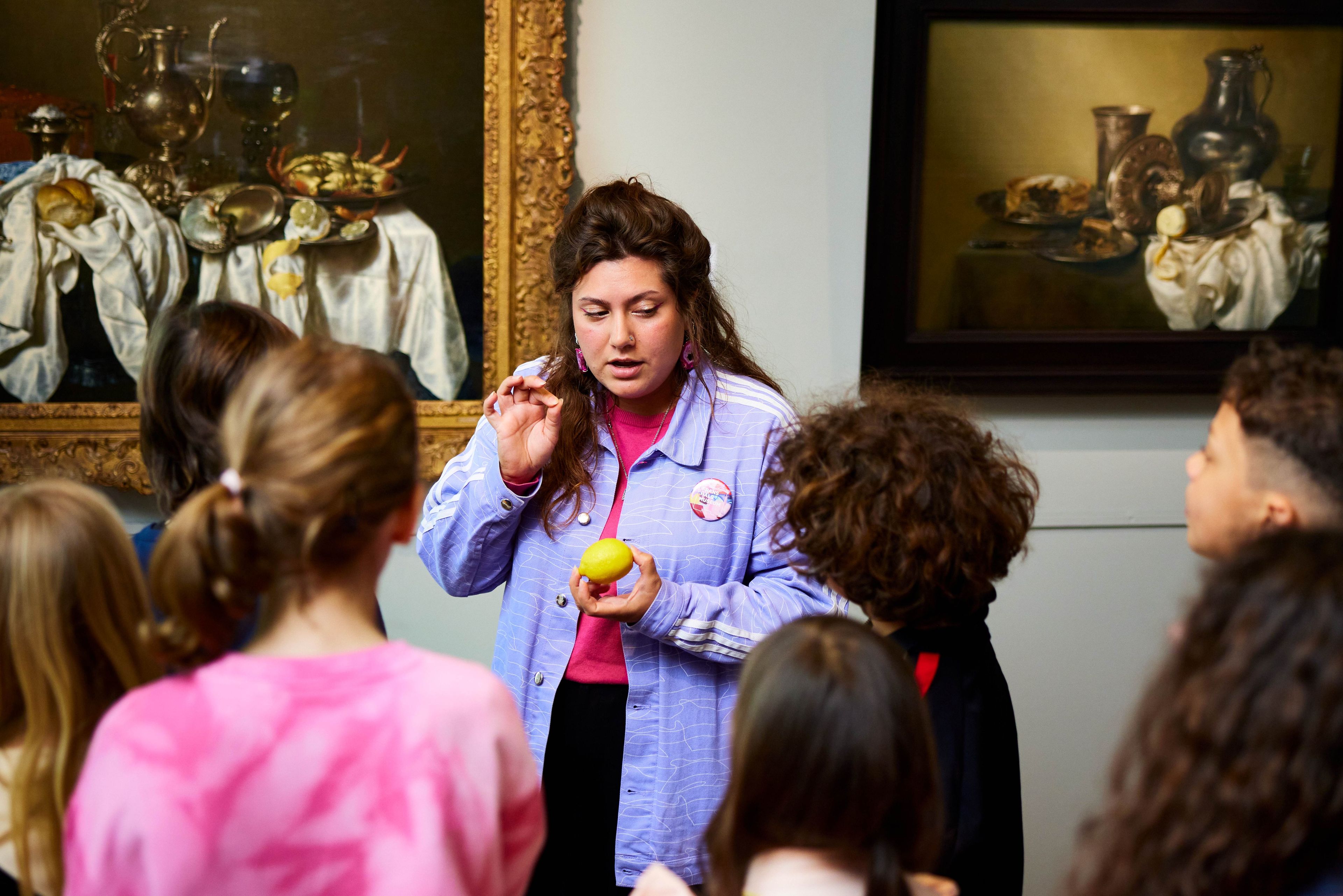 Museumdocent legt meer uit over twee stilleven schilderijen aan groep leerlingen.