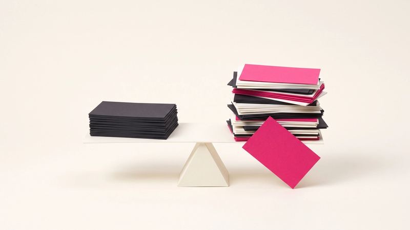 A paper balance scale with a stack of cards on one side and a taller, uneven stack on the other, one card slipping off