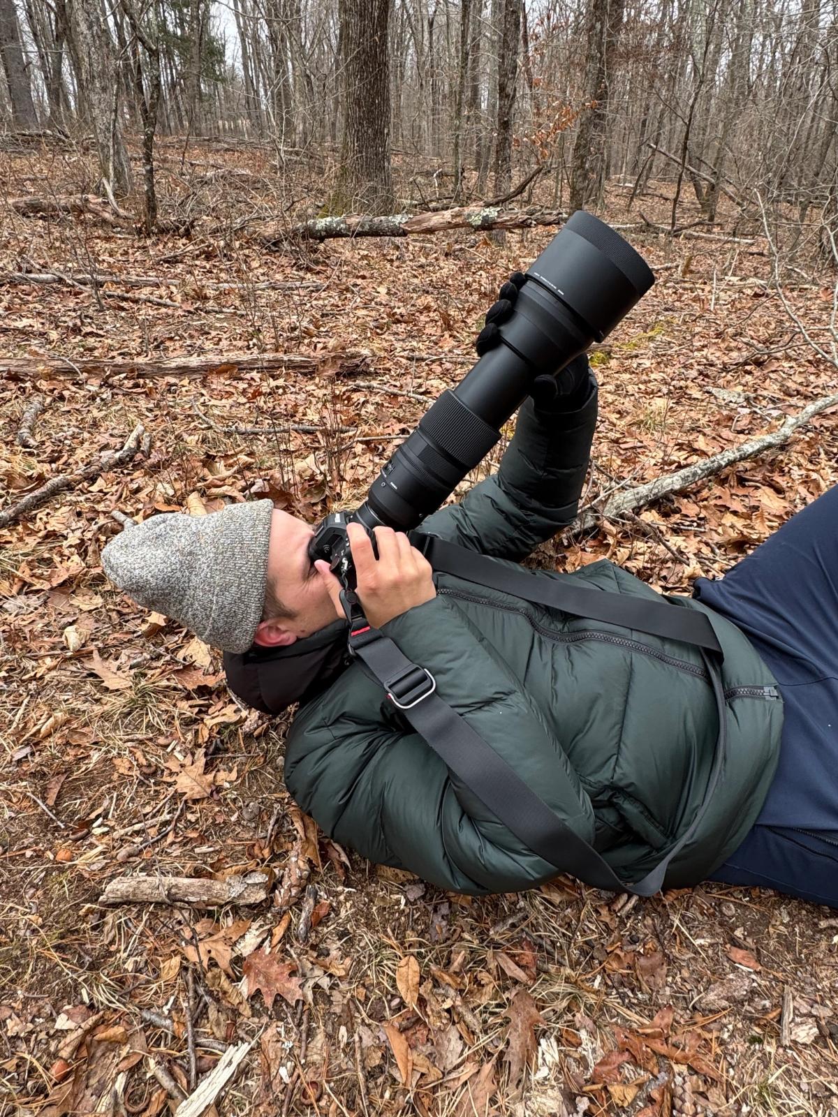 Ben lying on his back on a leaf-covered forest floor in a dark green jacket and grey knit hat, aiming a camera with a long telephoto lens up into the bare winter trees.