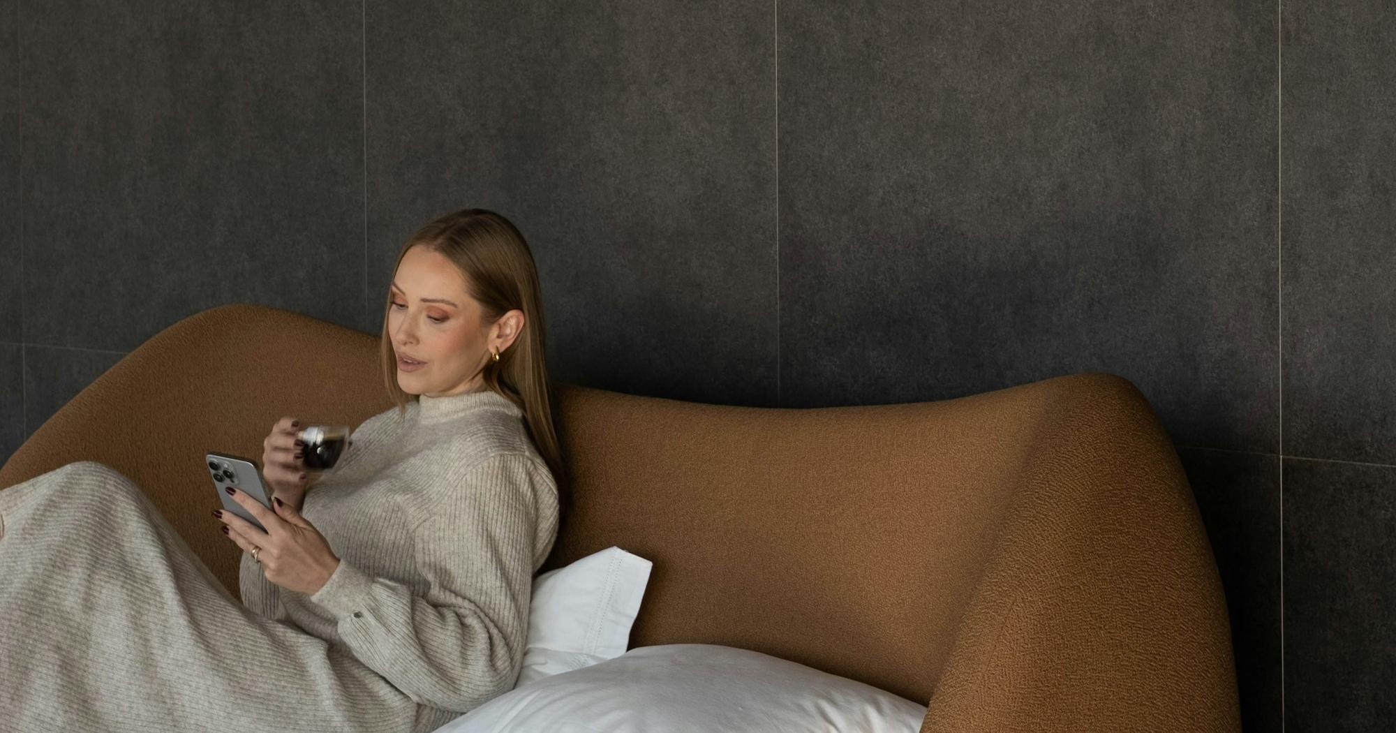 Woman sitting in bed in the morning with a mug of coffee, looking at her phone