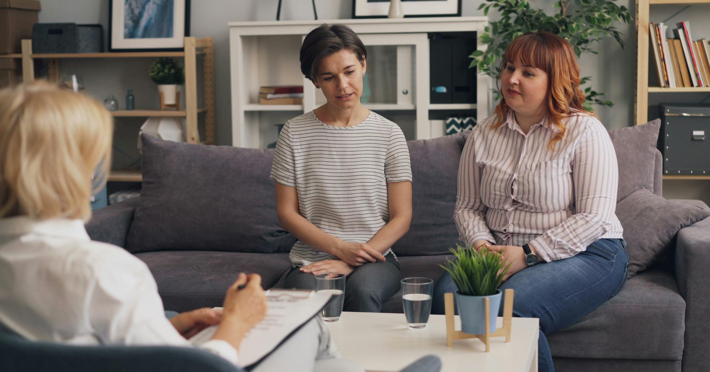 Two women sitting on a grey couch in a bright room, one listening closely as the other talks