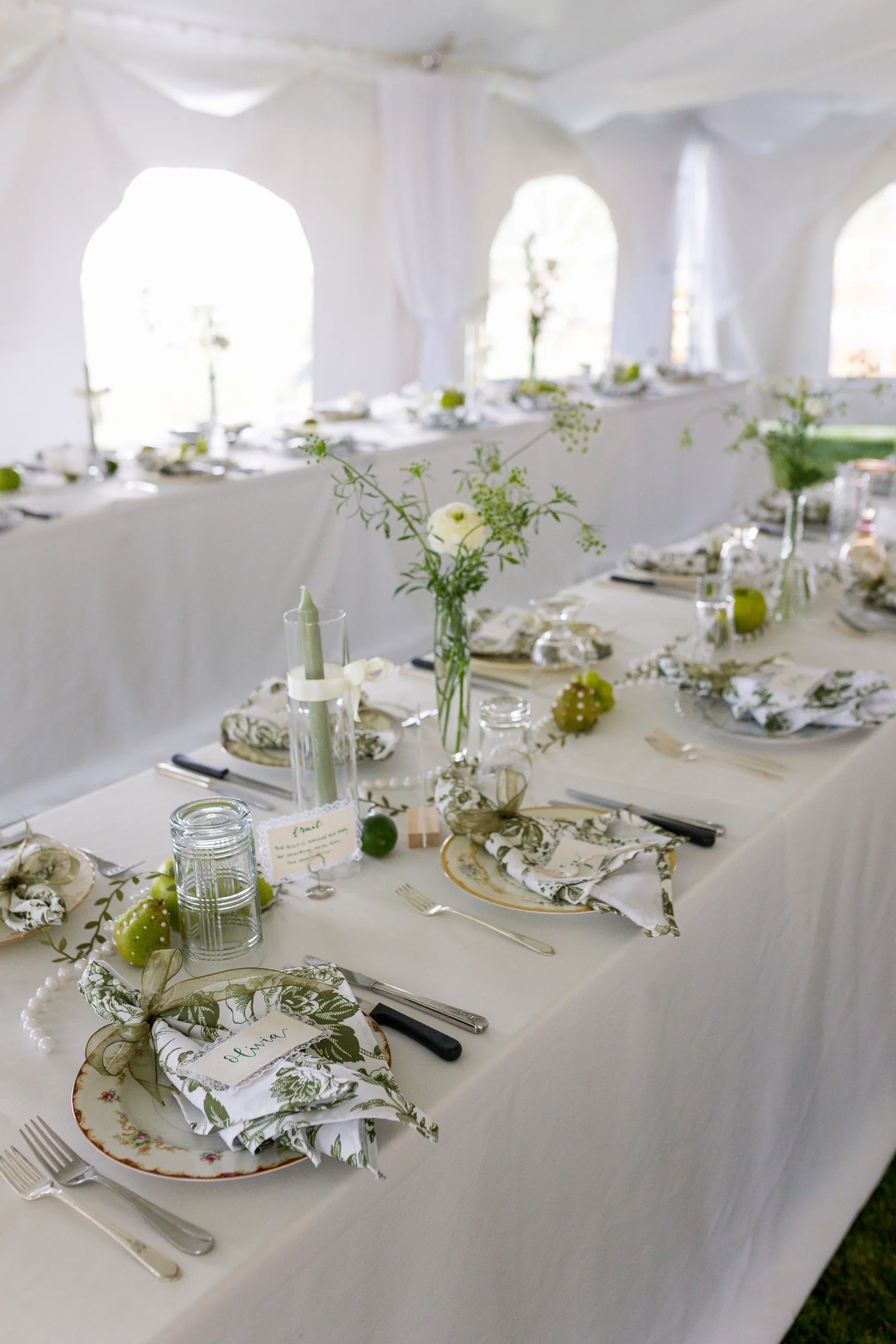 Long reception table under a white tent with a bud vase of white ranunculus and Queen Anne's lace, green toile napkins and a trail of green fruit and pearls
