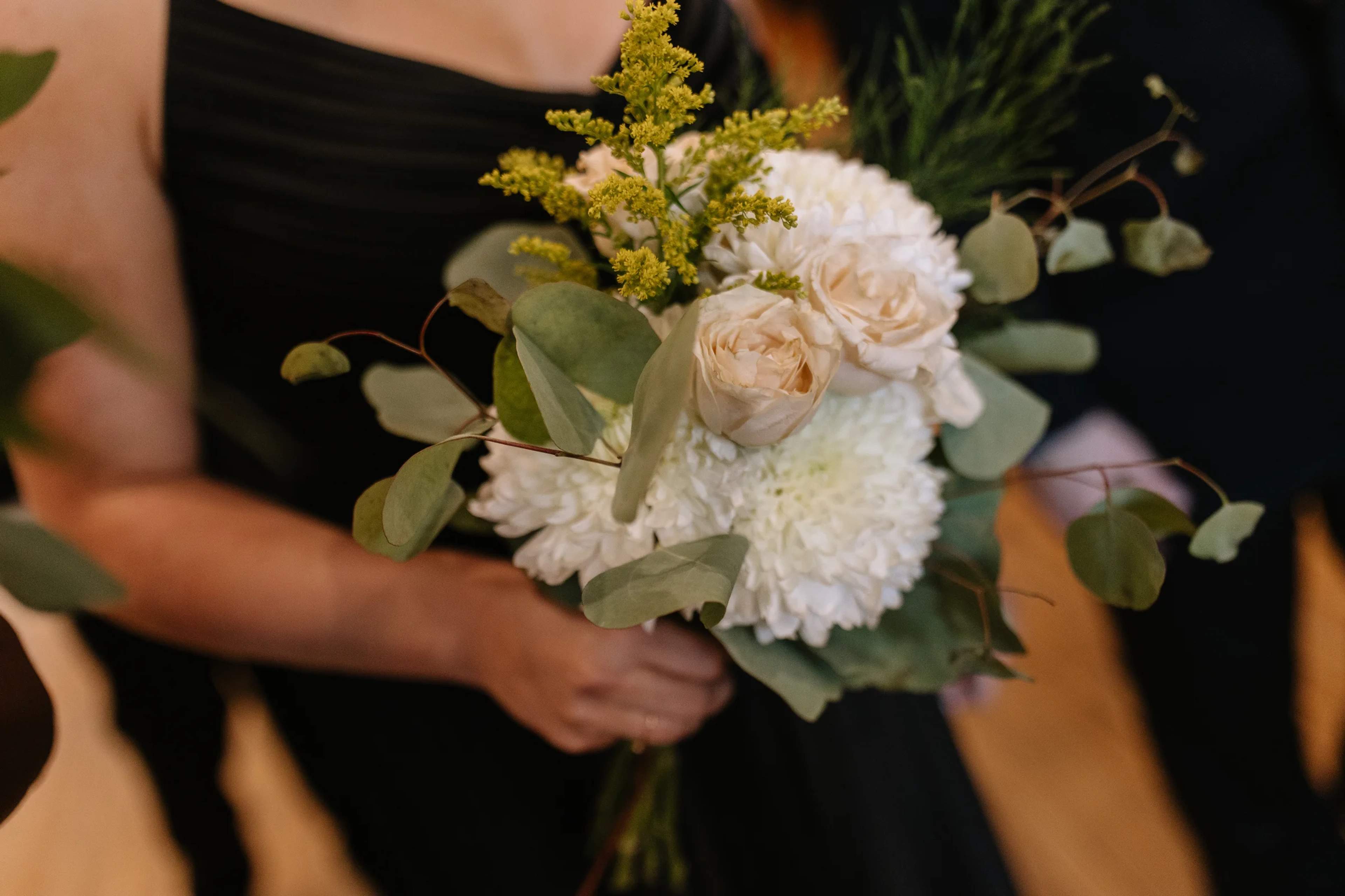 Close-up of a bridesmaid bouquet of white roses, white mums, goldenrod and silver dollar eucalyptus held against a black gown