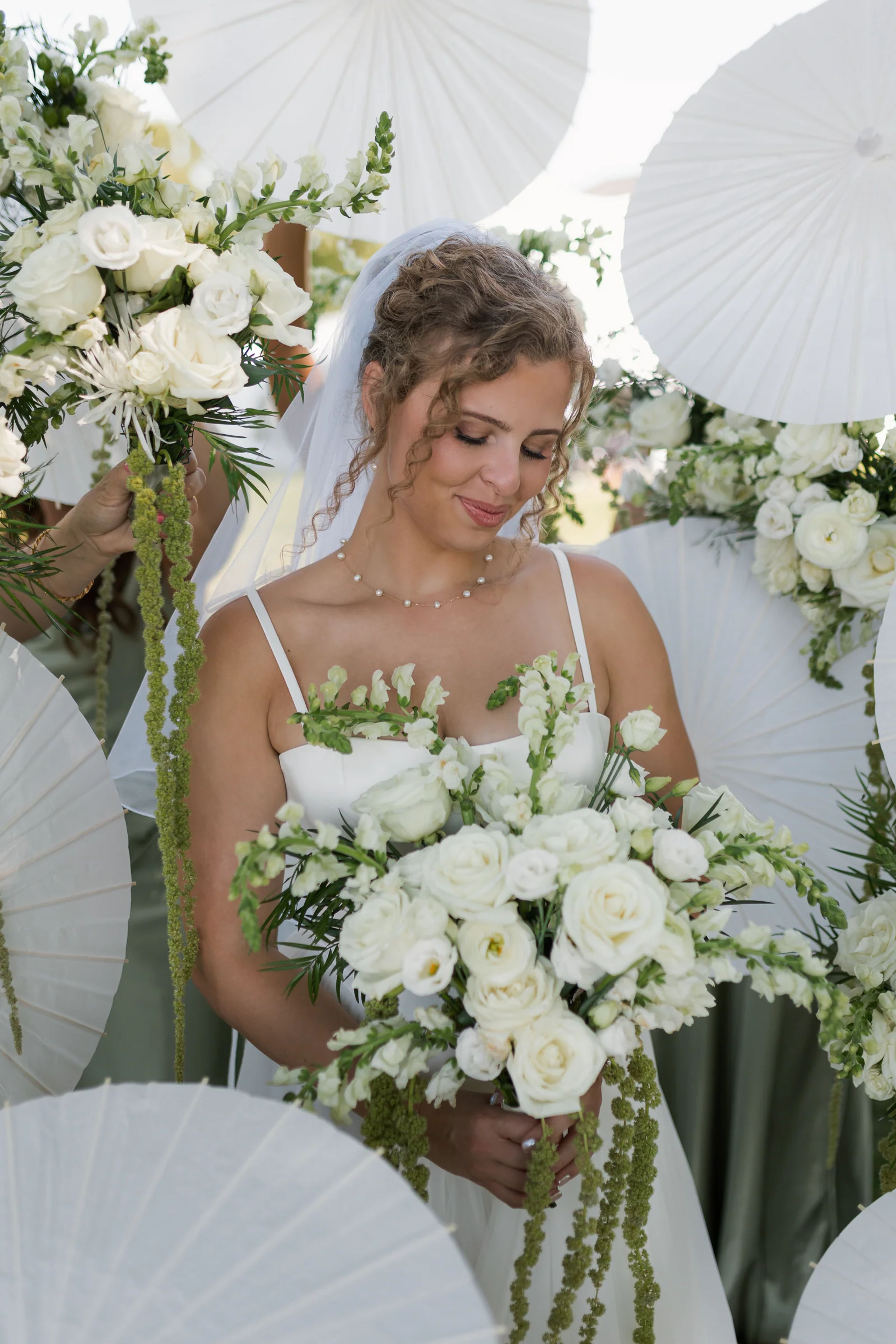 Bride looking down at a bouquet of white ranunculus, garden roses and snapdragons with trailing green amaranthus, framed by white paper parasols