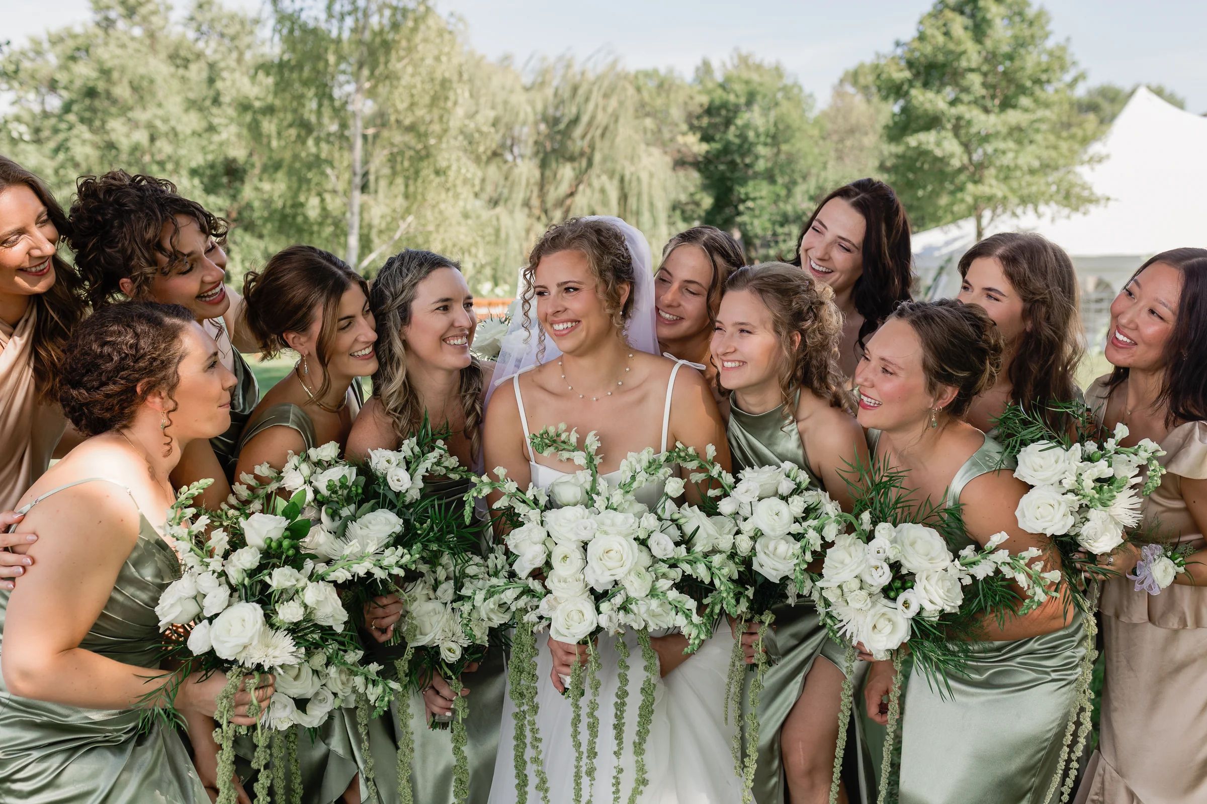 Bride and bridesmaids in sage silk laughing over a row of white rose and ranunculus bouquets with trailing amaranthus