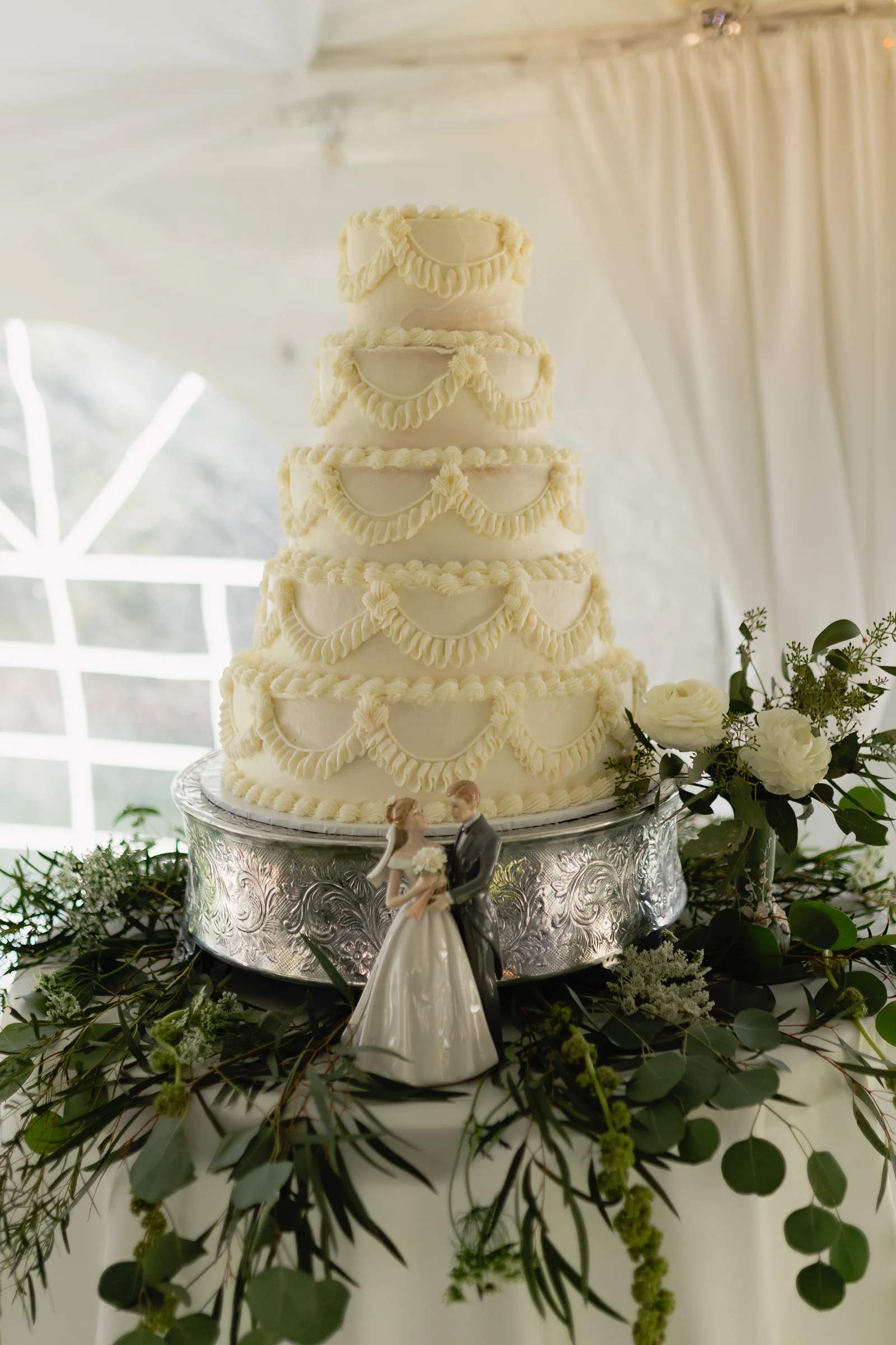 Five-tier piped white cake on a silver stand ringed with eucalyptus, white ranunculus and trailing green amaranthus
