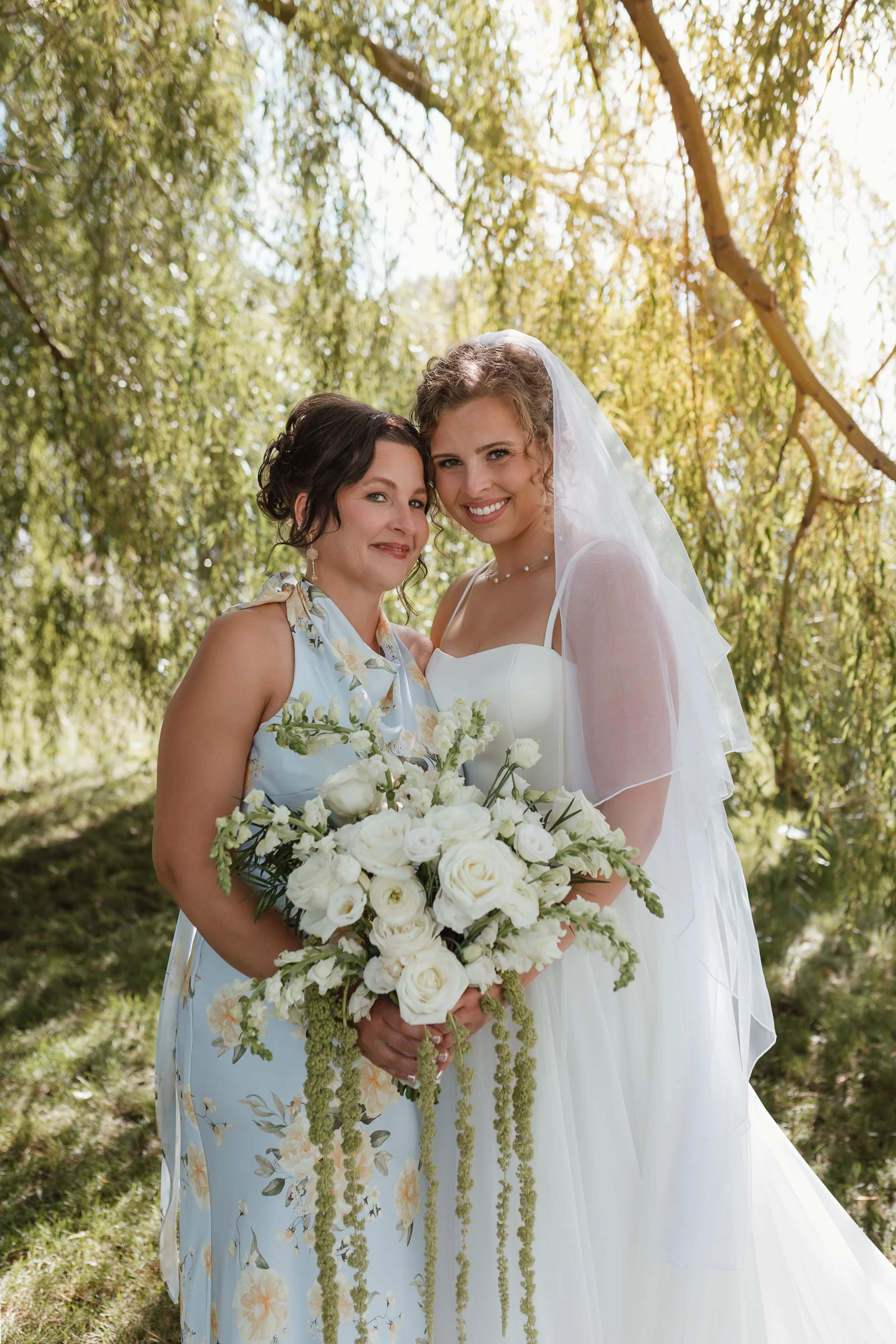 Bride beside a guest in a pale blue floral dress under a willow, holding a white rose, ranunculus and snapdragon bouquet with trailing amaranthus