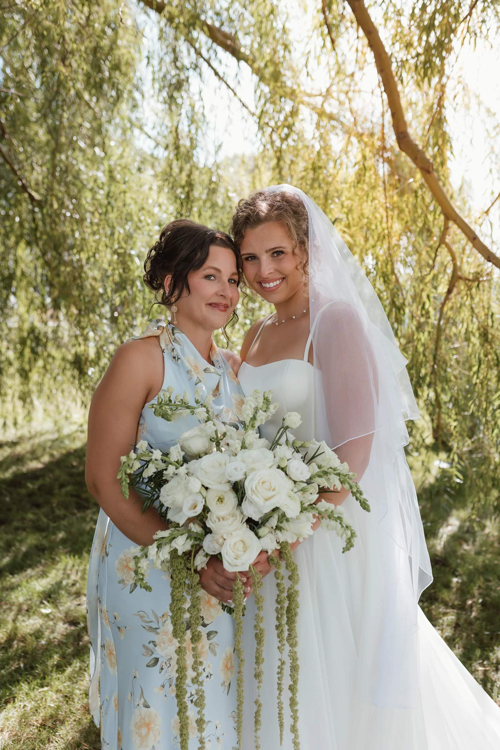 Bride beside a guest in a pale blue floral dress under a willow, holding a white rose, ranunculus and snapdragon bouquet with trailing amaranthus