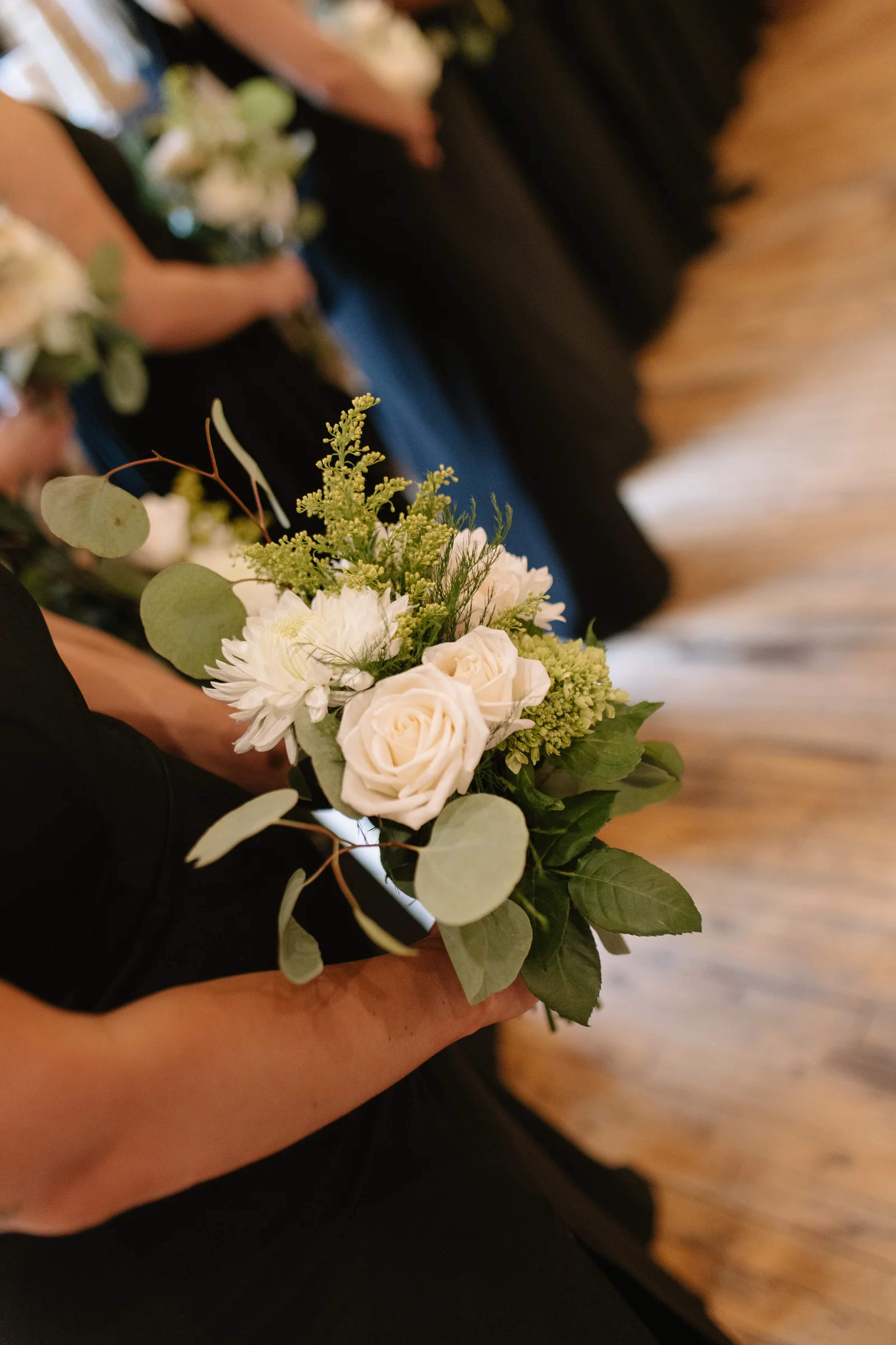 Bridesmaid bouquet of white roses, a white mum, goldenrod and eucalyptus in the foreground, with a line of bridesmaids receding across a wood floor