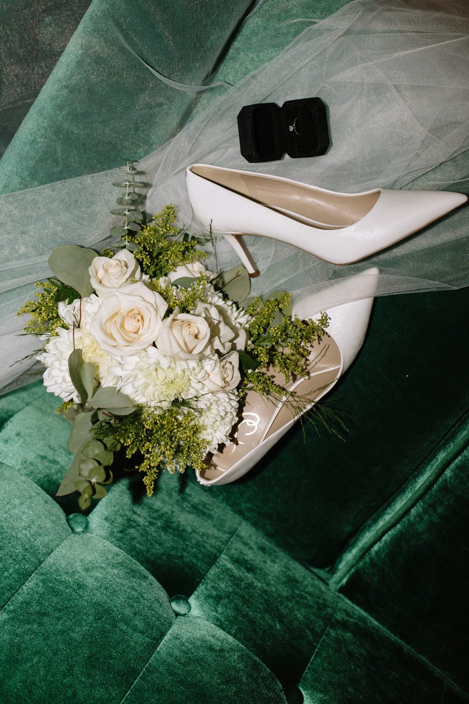 Flat lay of a bridal bouquet of white roses, football mums, goldenrod and silver dollar eucalyptus beside white pointed pumps, a tulle veil and a ring box on green velvet