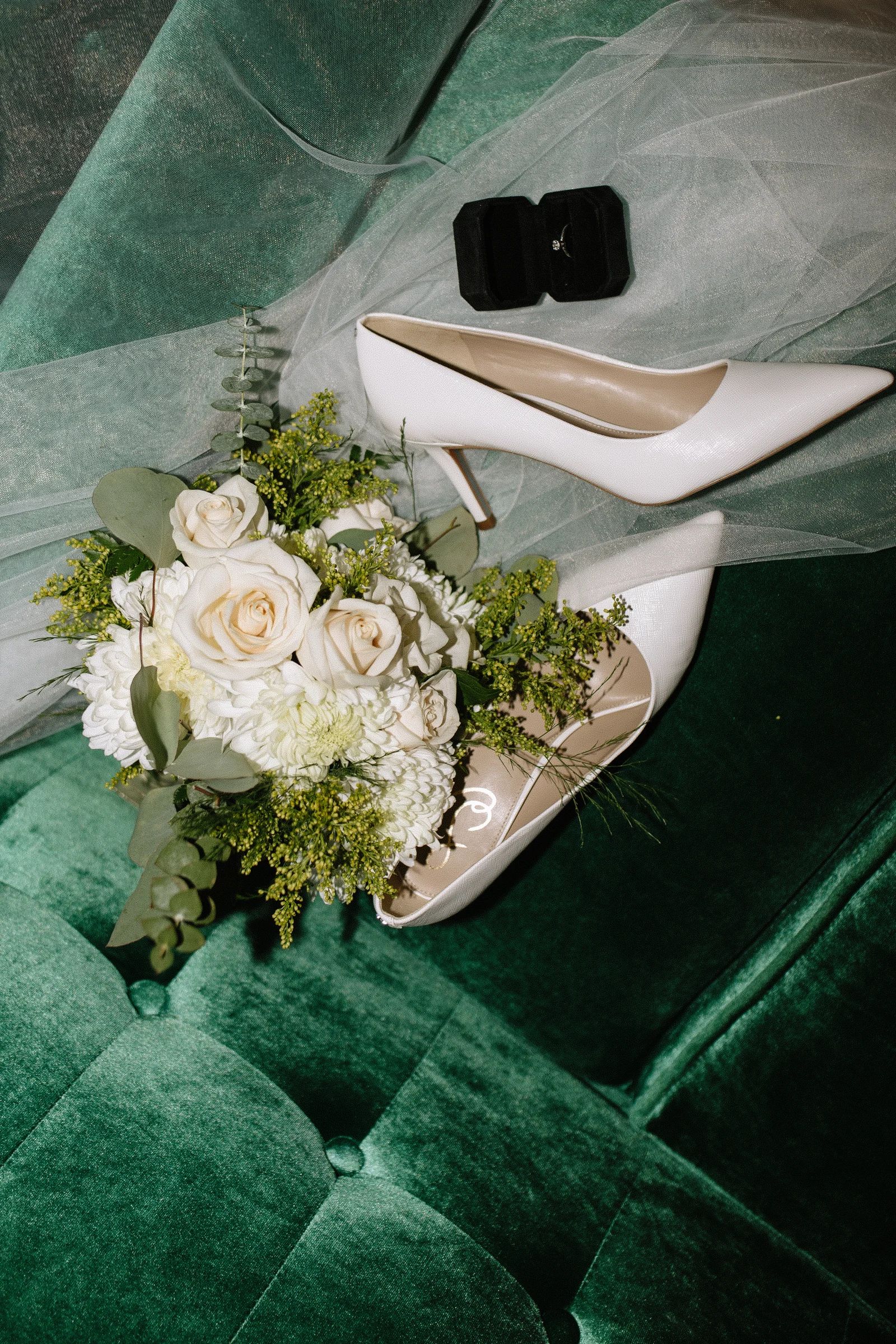 Flat lay of a bridal bouquet of white roses, football mums, goldenrod and silver dollar eucalyptus beside white pointed pumps, a tulle veil and a ring box on green velvet