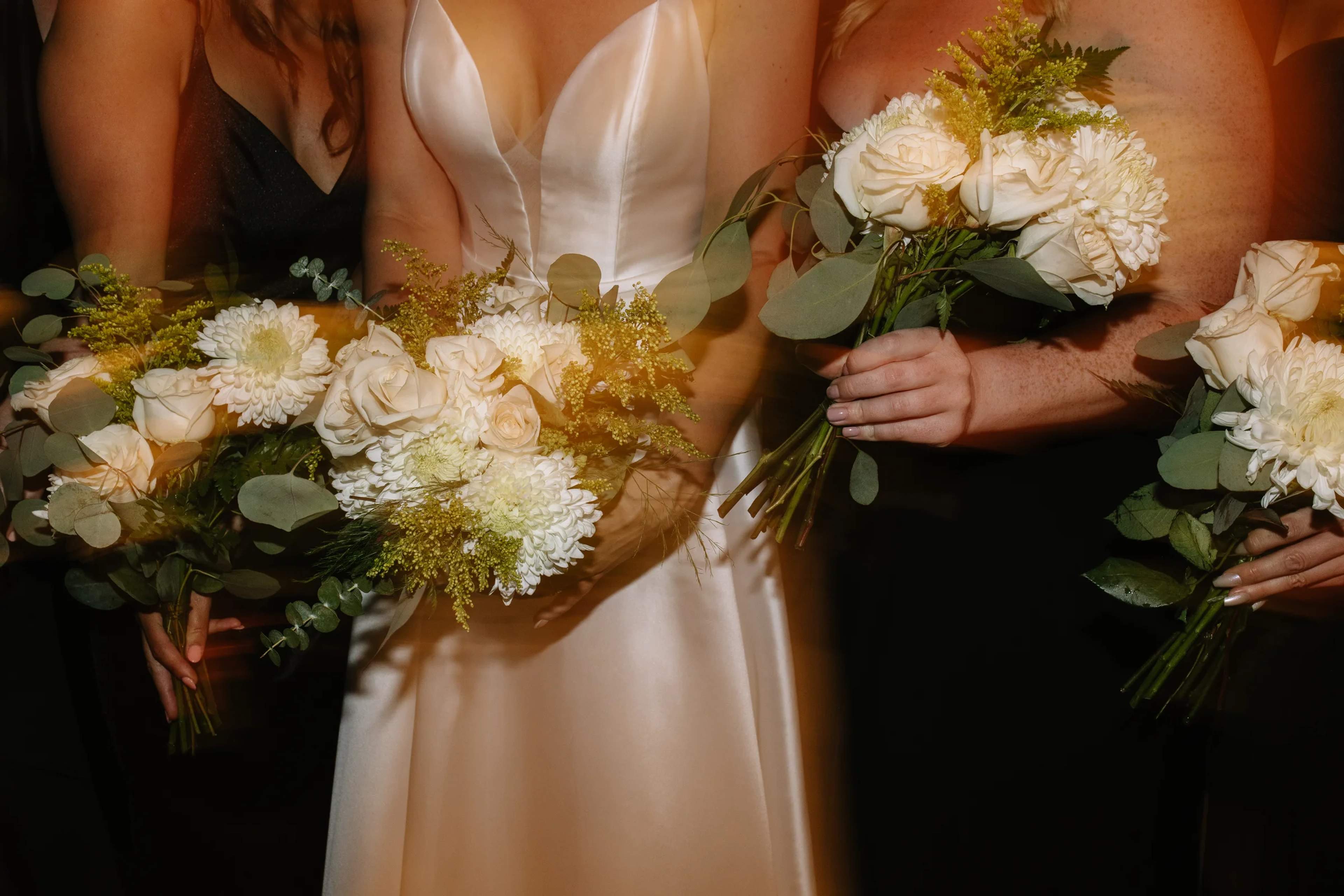 Bride and bridesmaids in black holding bouquets of white roses, mums, goldenrod and eucalyptus, lit by warm flash