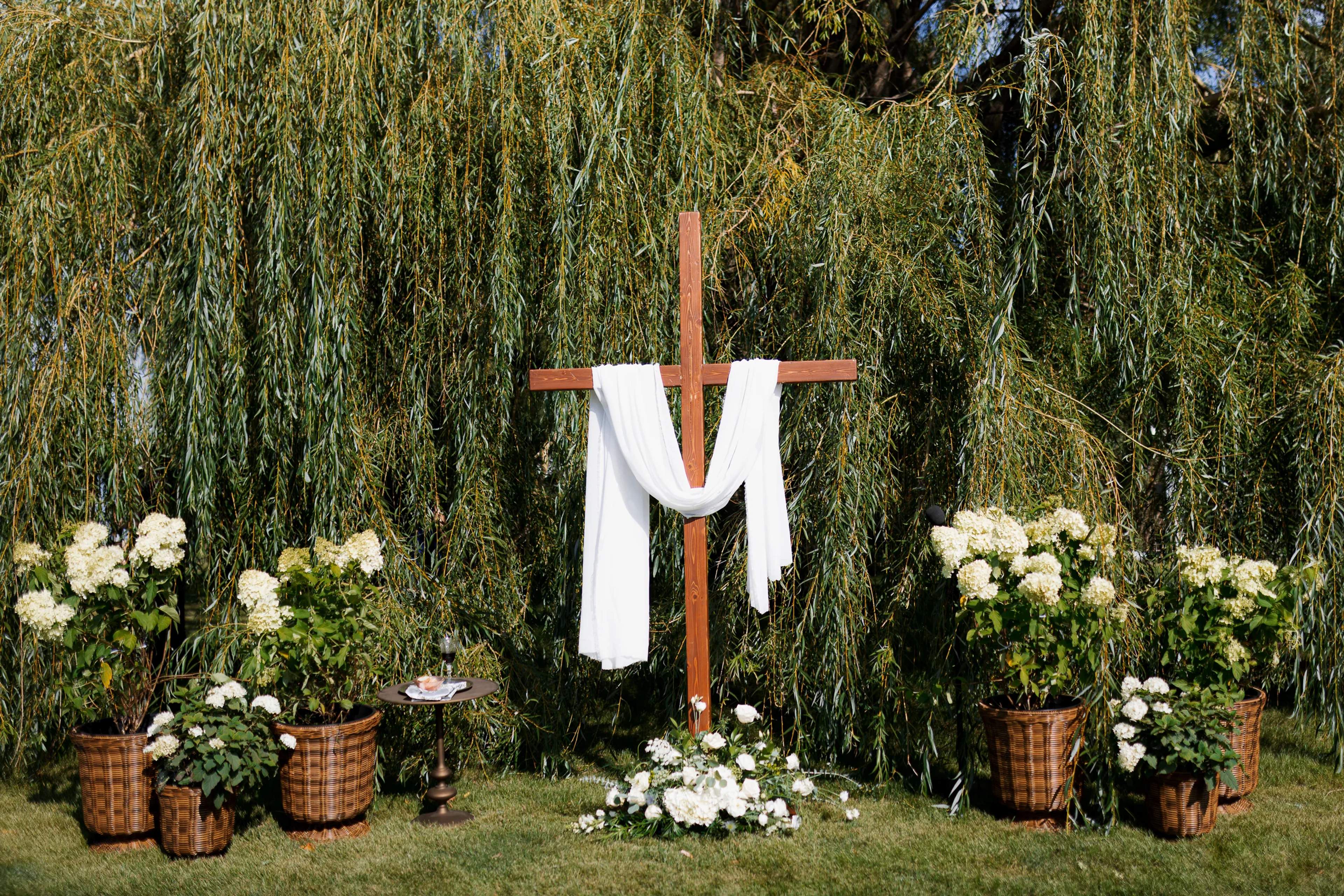 Wooden cross draped in white fabric beneath a weeping willow, flanked by basket planters of white hydrangea and a low meadow of white roses and hydrangea at its base