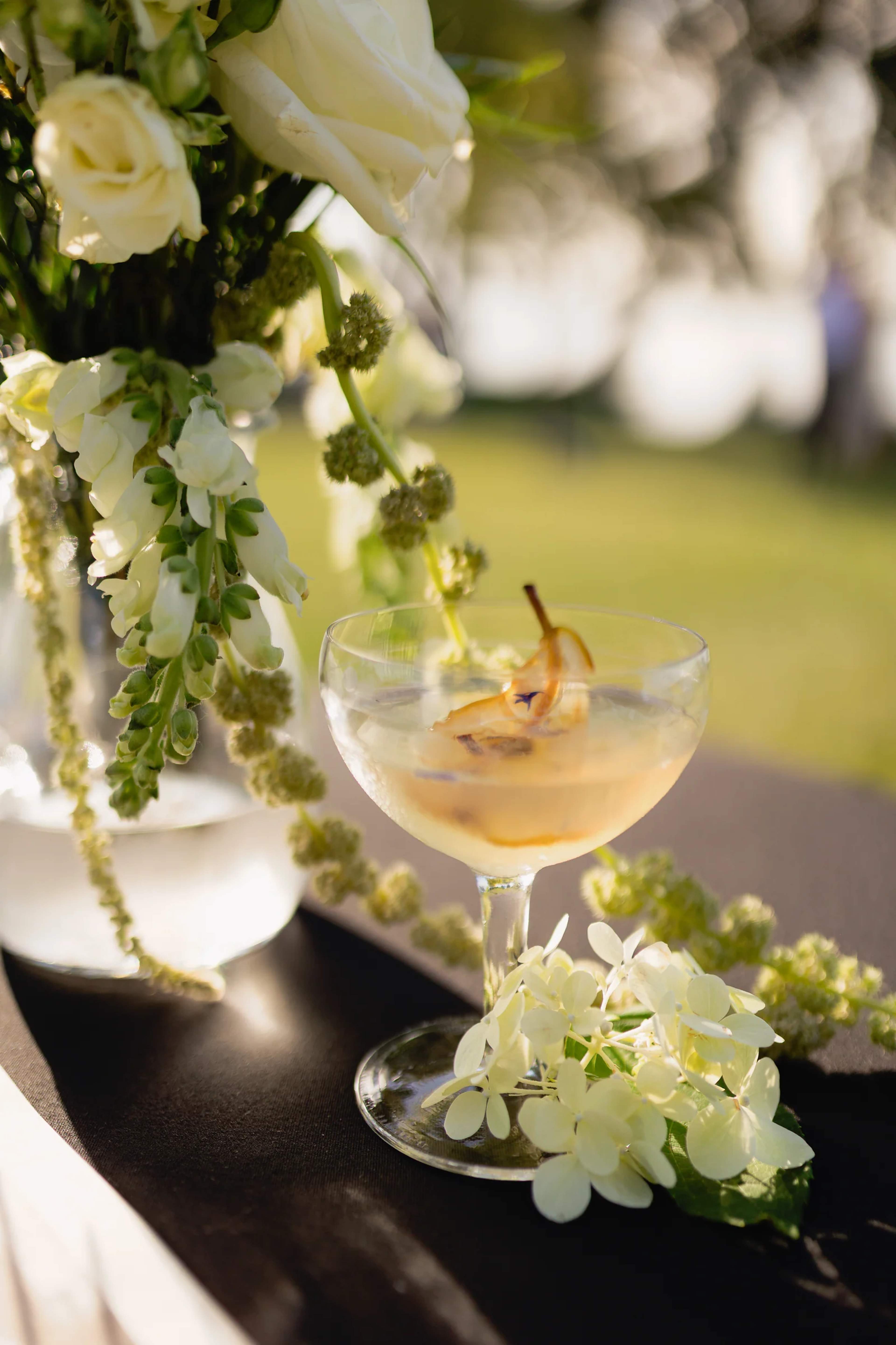 Coupe cocktail with a poached pear beside a vase of white roses and snapdragons, trailing amaranthus and a sprig of white hydrangea on a dark bar