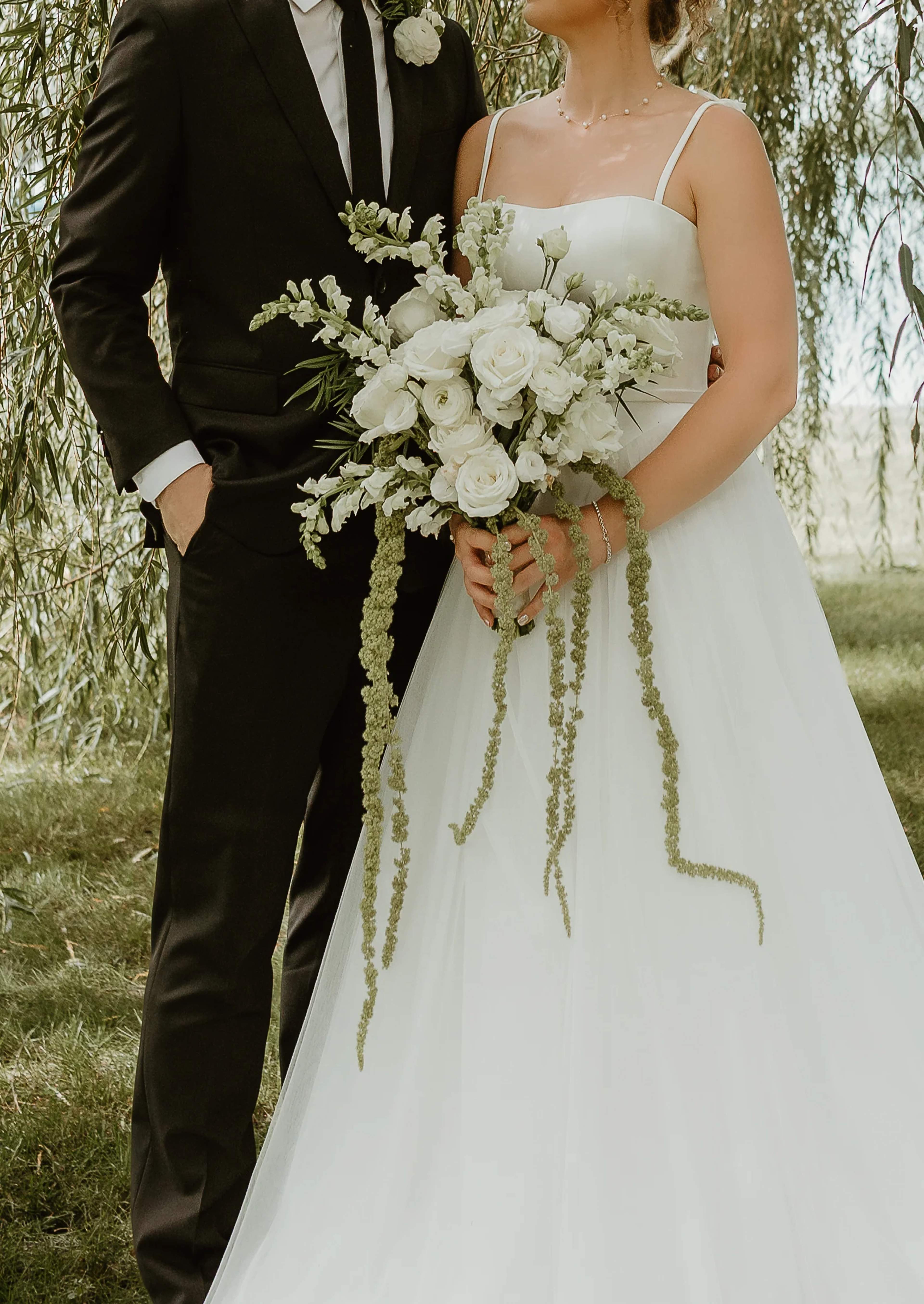 Bridal bouquet of white roses, ranunculus and snapdragons with long trailing amaranthus, held between an ivory gown and a black suit with a single white boutonniere