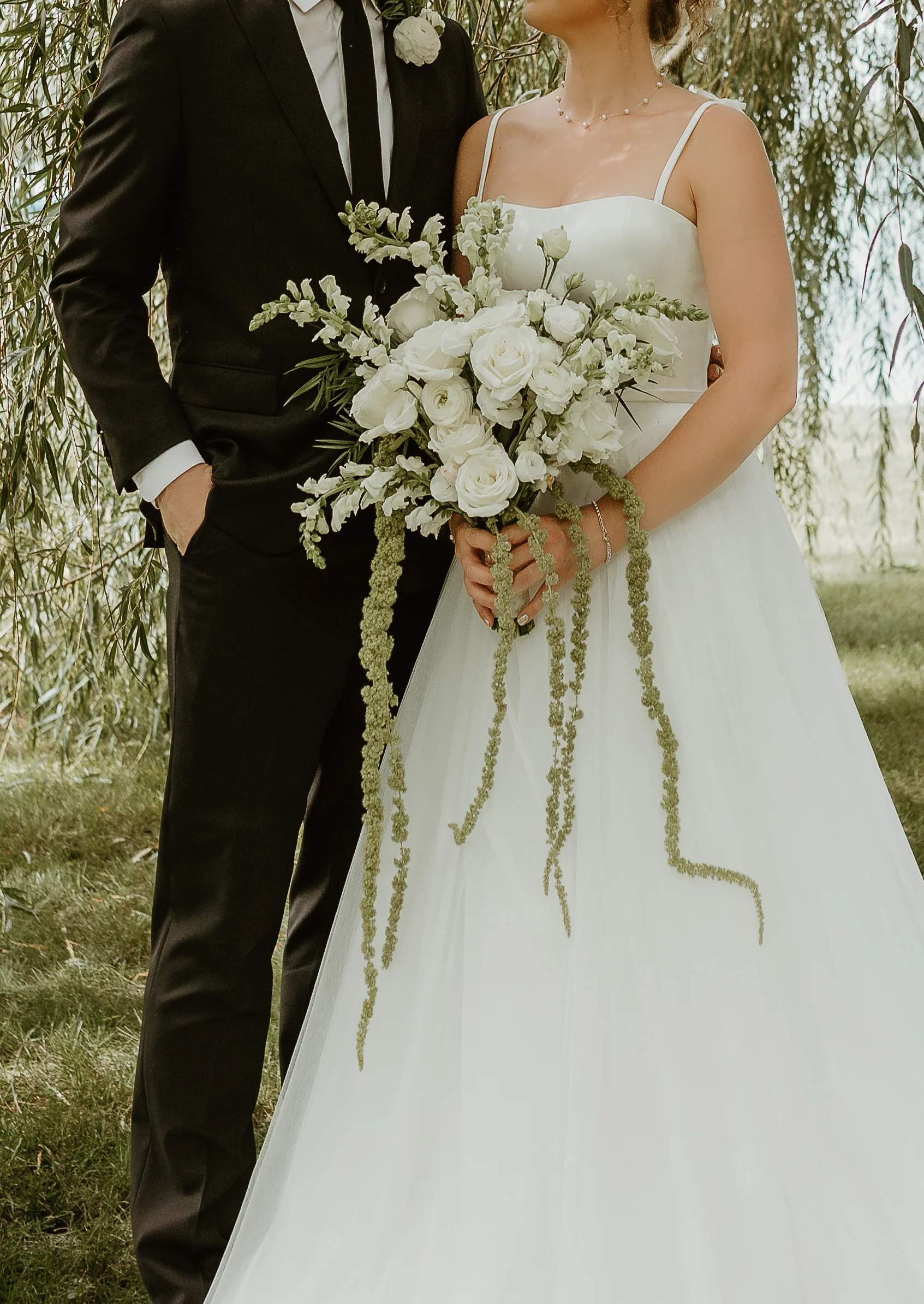 Bridal bouquet of white roses, ranunculus and snapdragons with long trailing amaranthus, held between an ivory gown and a black suit with a single white boutonniere