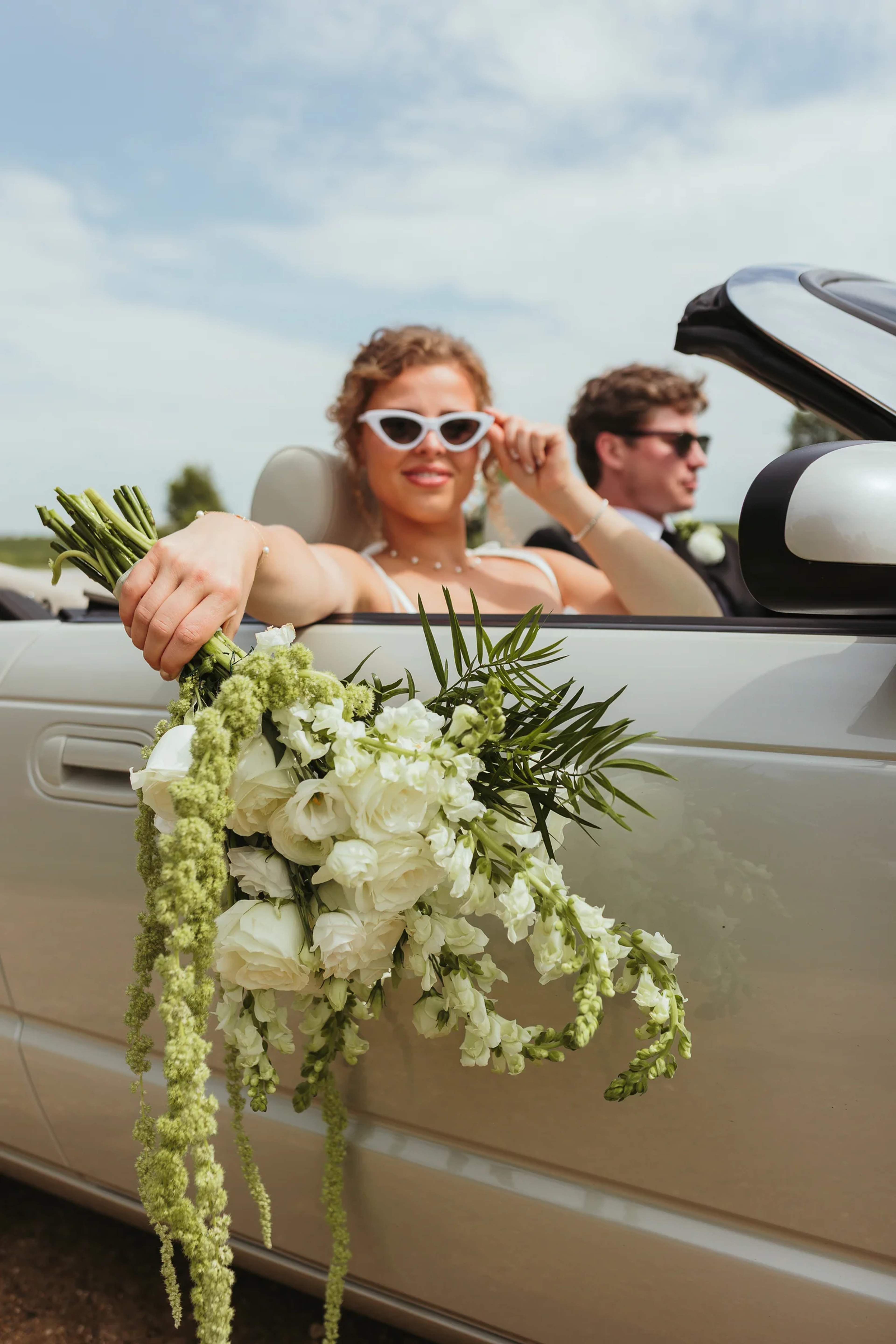 Bride in white sunglasses leaning out of a vintage convertible, holding a white rose and snapdragon bouquet with trailing amaranthus and a palm frond