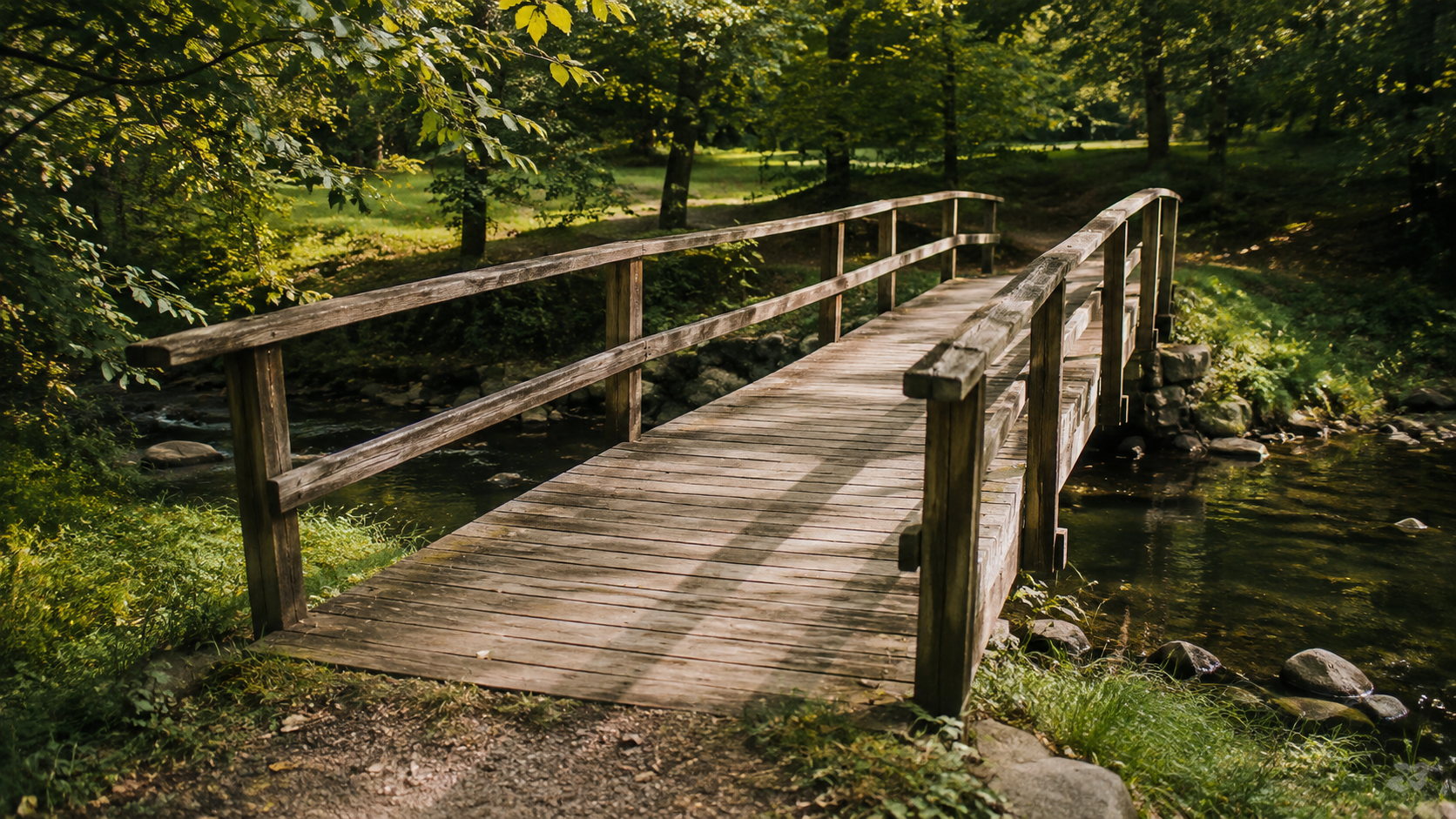 A wooden footbridge with railings crosses a stream beneath green trees.