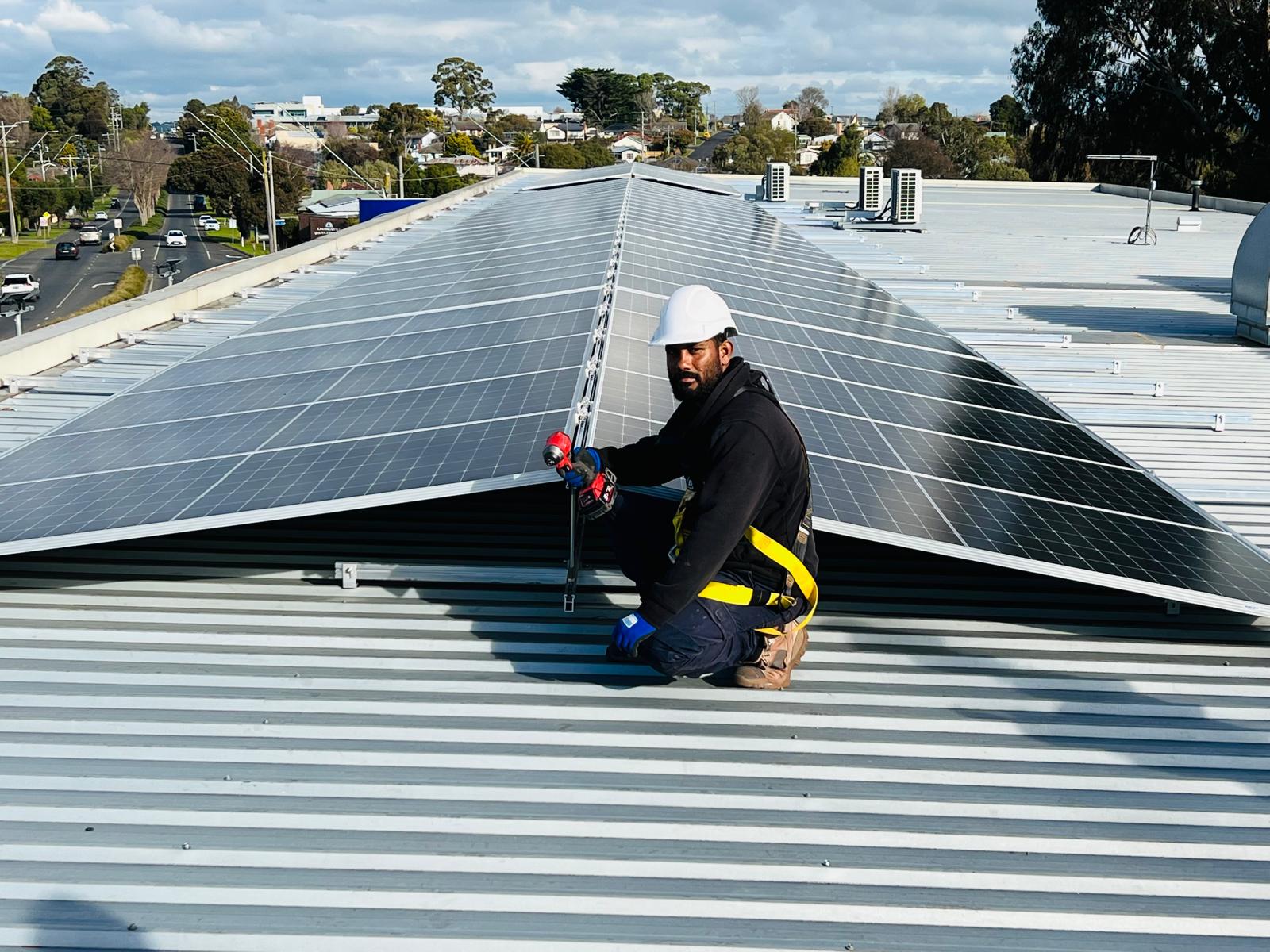 A JM Solar installer in a hard hat and safety harness fixing panels on a metal roof