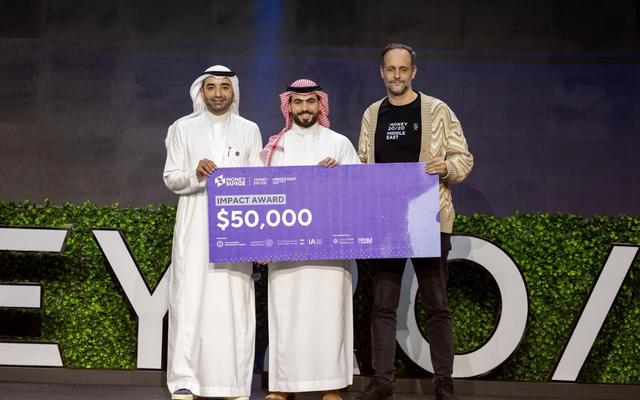 Three men stand on the Money20/20 Middle East stage; the Eddekhar representative in the center holds a purple "Impact Award $50,000" plaque, flanked by two colleagues, with the Money20/20 backdrop behind them.
