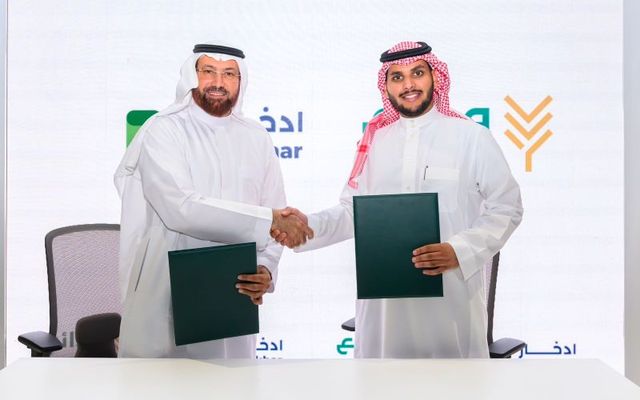 Dr. Adnan Alkhiary, CEO of Eddekhar, and Abdullah Alandas, CEO of Wadaie, shake hands after signing their partnership agreement, each holding a green signed document, with the Eddekhar and Wadaie logos on the backdrop behind them.