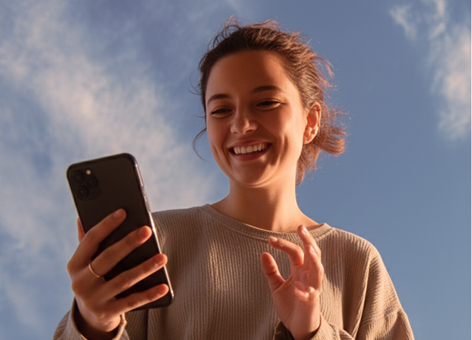 Woman smiling while holding smartphone under a sunny blue sky. Social media, connection, joyful.