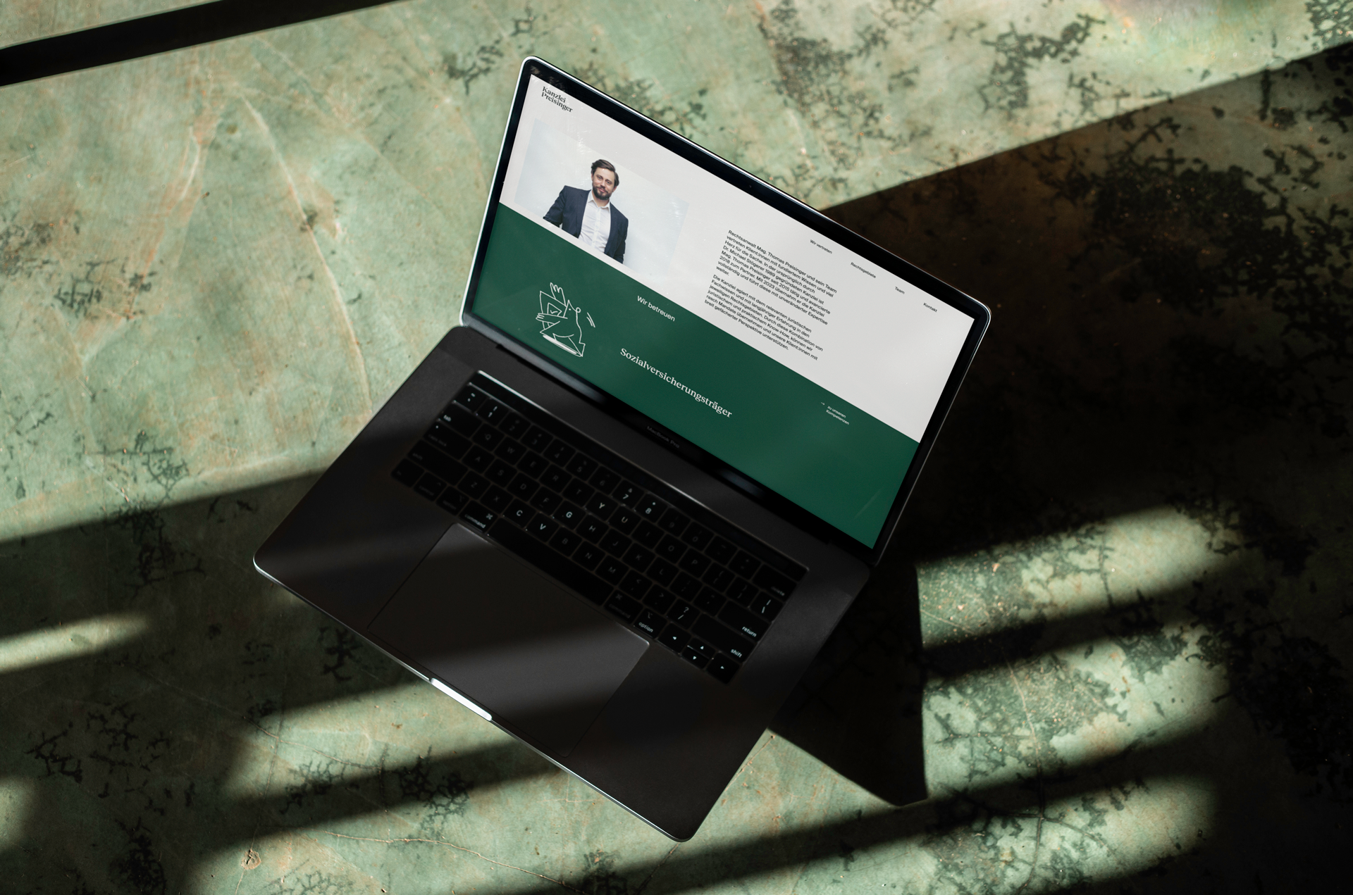 Ein Laptop auf einem grünen, strukturierten Untergrund zeigt die Website der Kanzlei Preisinger. Die Website hat eine professionelle Gestaltung mit einem Bild eines Anwalts im oberen Bereich und Textinformationen daneben. Der untere Bereich ist grün und enthält weitere Informationen über die Dienstleistungen der Kanzlei.