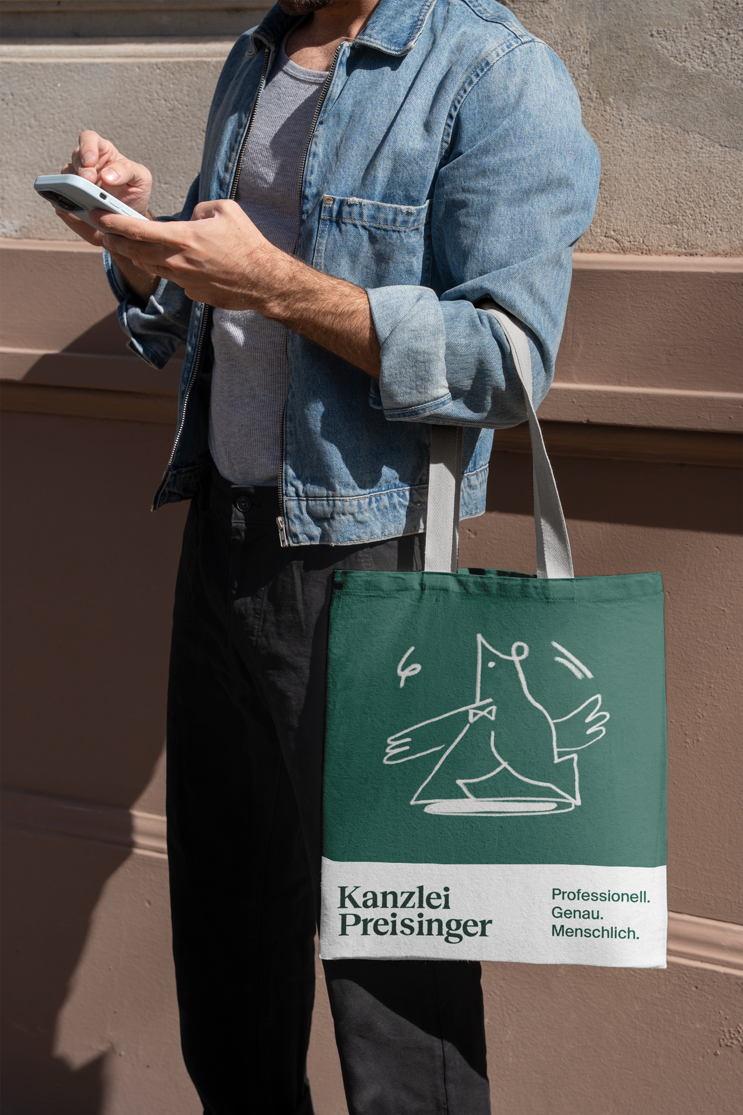 Ein Mann in einer Jeansjacke und einem grauen T-Shirt hält ein Smartphone in der Hand und trägt eine grüne Stofftasche mit dem Logo der