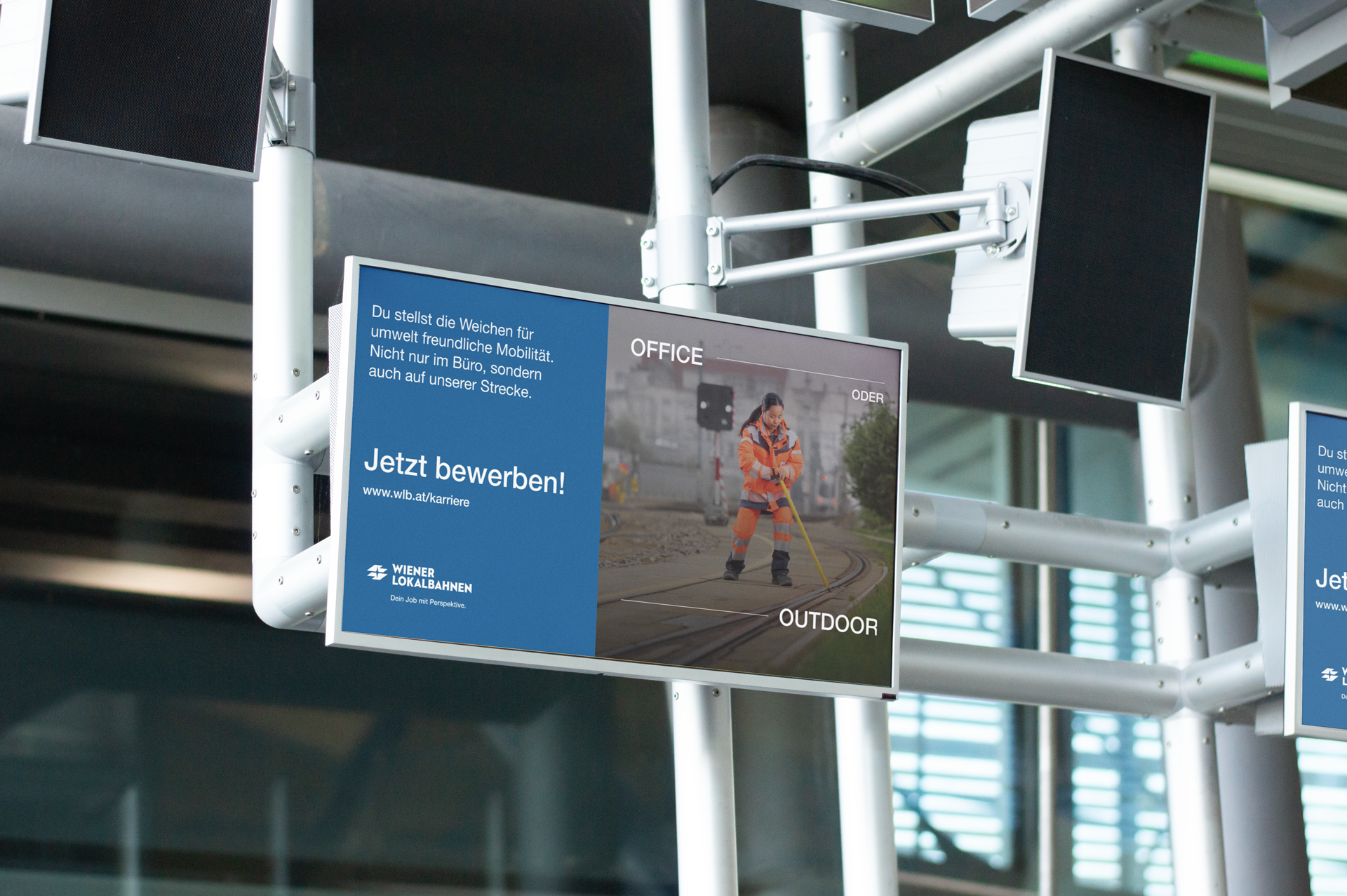 Das Bild zeigt ein digitales Reklameschild in einem Bahnhofsgebäude. Auf dem Bildschirm ist eine Stellenanzeige der Wiener Lokalbahnen zu sehen. Der Text auf der linken Seite des Bildschirms lautet: