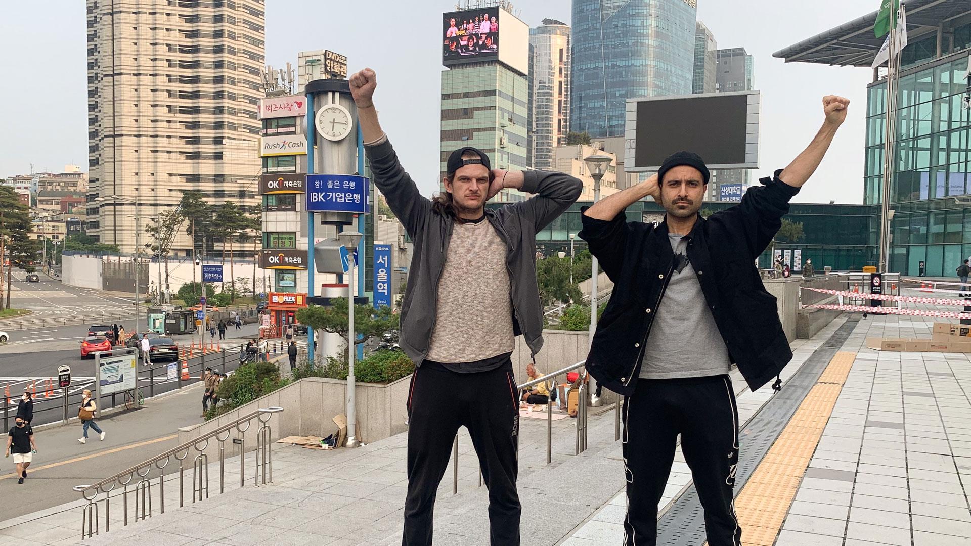 Two men on a city street making the Deaf Power sign: one palm over an ear, the other fist raised