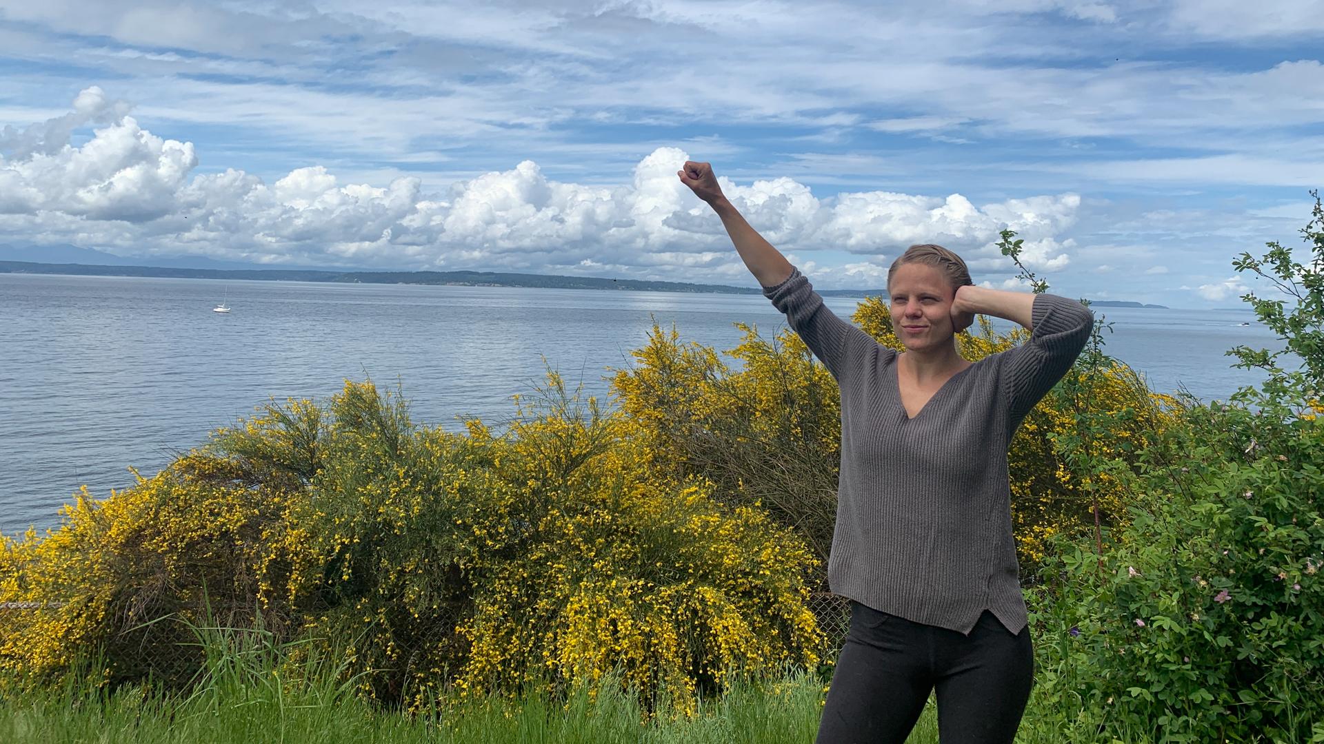 A woman by the sea among yellow gorse making the Deaf Power sign