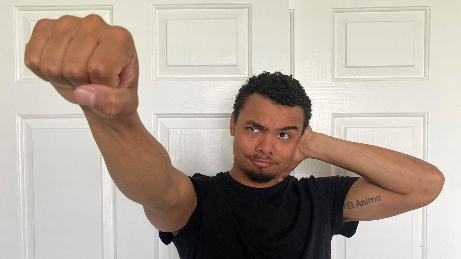 A man in front of a white door making the Deaf Power sign, fist toward the camera