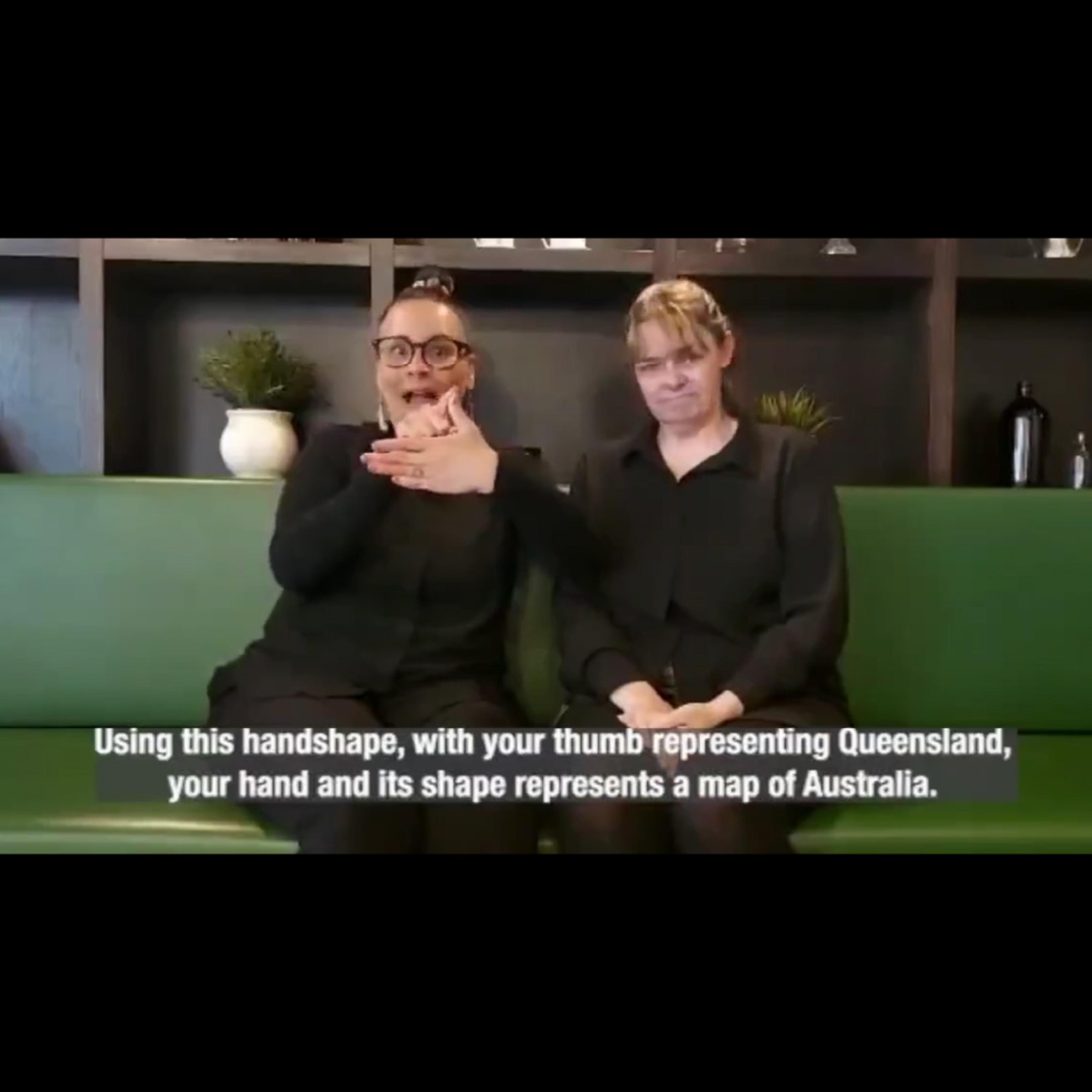 Two women signing on a green sofa, captioned about the handshape for Queensland