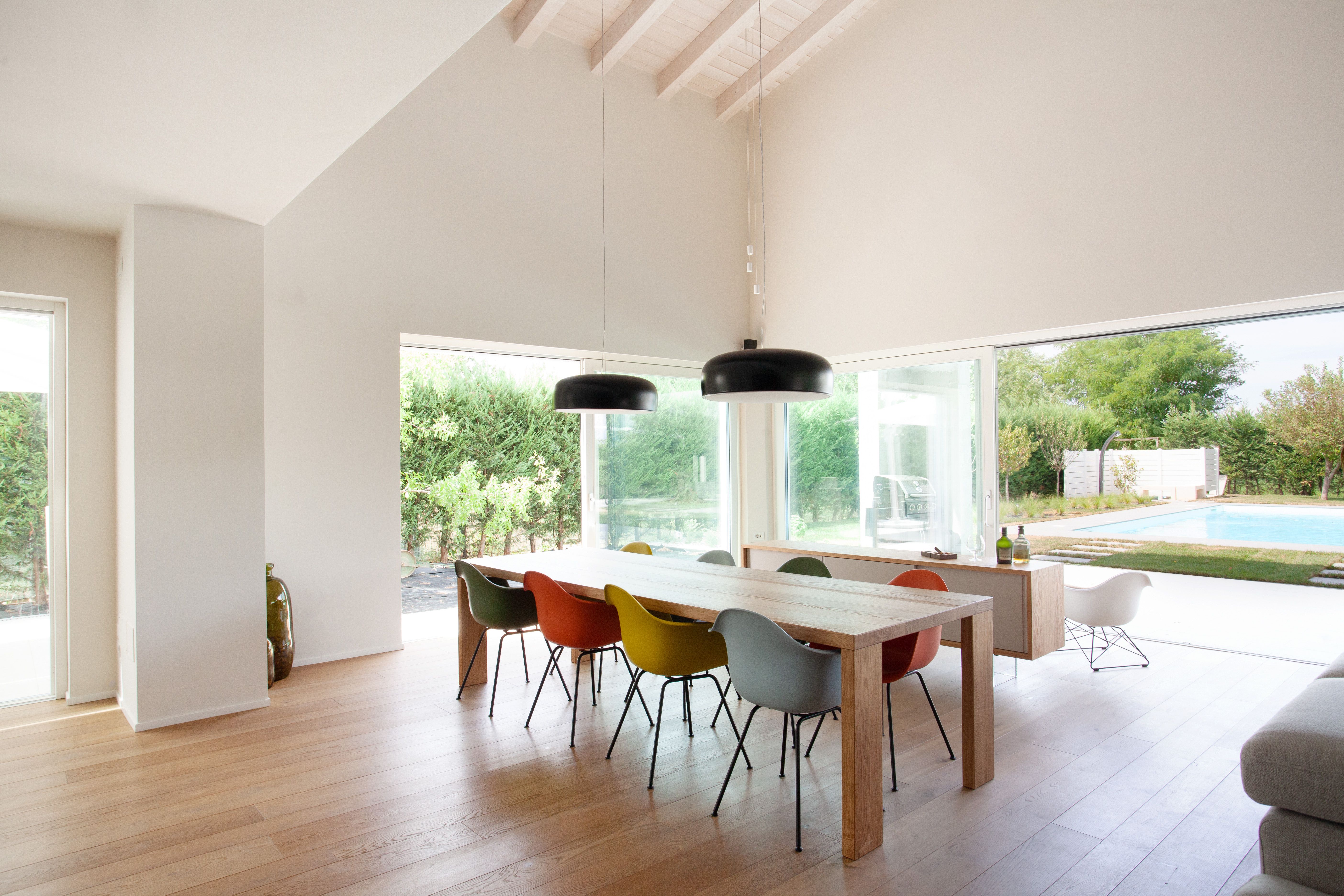 immagine a colori con un grande tavolo da pranzo, con delle sedie colorate e una piscina che si vede esternamente.
