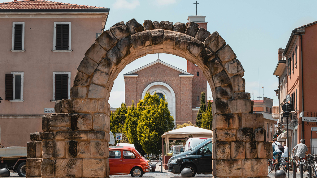 A stone arch frames a street scene with buildings, cars, and people.