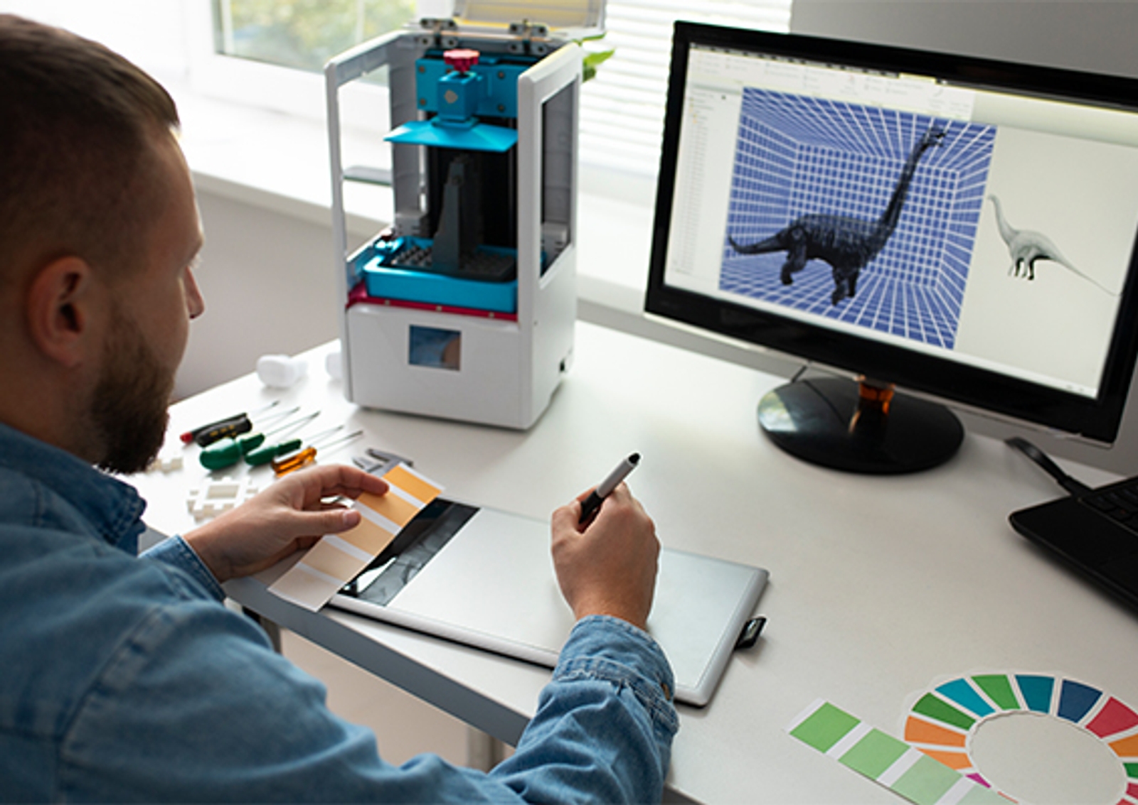 Photo d'un designer examinant un modèle 3D de dinosaure sur un écran d'ordinateur, à côté d'une imprimante 3D de bureau