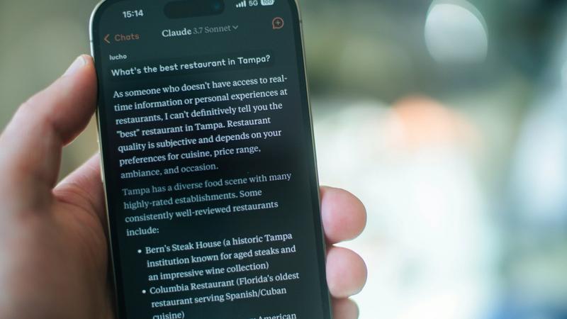 A hand holding a smartphone displaying a Claude AI conversation answering the question "What's the best restaurant in Tampa?" — illustrating how consumers now use AI assistants to find local businesses