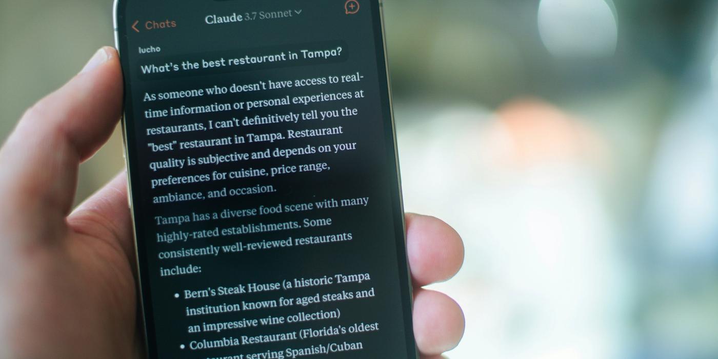 A hand holding a smartphone displaying a Claude AI conversation answering the question "What's the best restaurant in Tampa?" — illustrating how consumers now use AI assistants to find local businesses