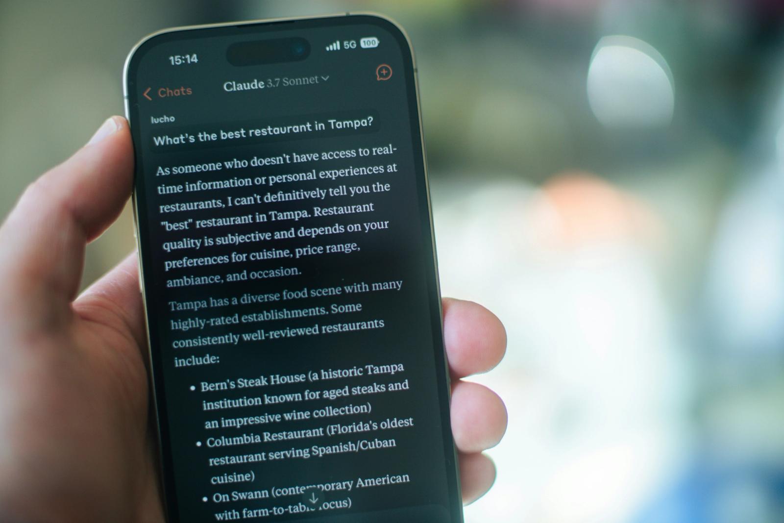 A hand holding a smartphone displaying a Claude AI conversation answering the question "What's the best restaurant in Tampa?" — illustrating how consumers now use AI assistants to find local businesses