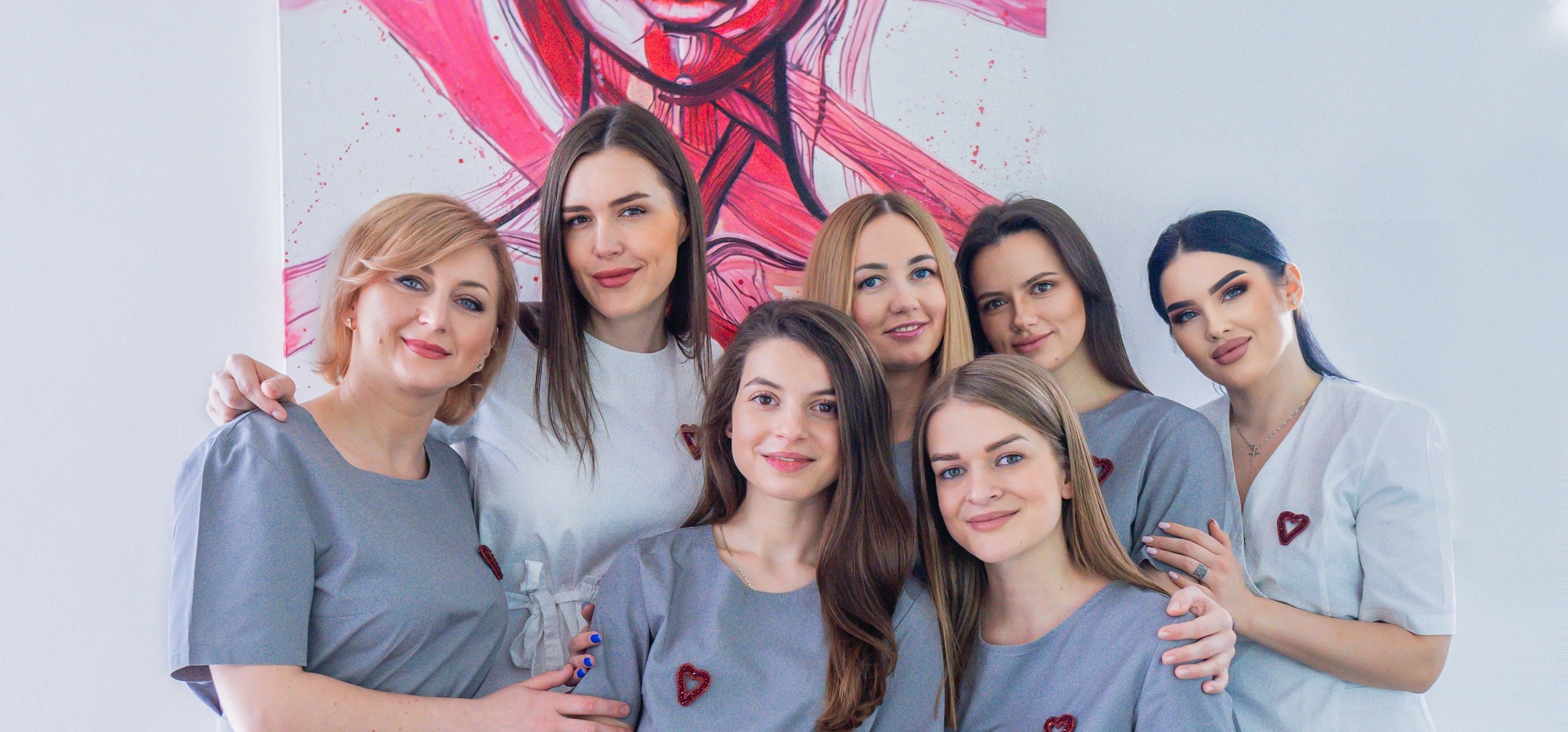 Група спеціалістів косметологічного центру, стоячи в їхньому робочому просторі. Вони усміхаються та виглядають дружелюбно, одягнені в професійний одяг з логотипами клініки.