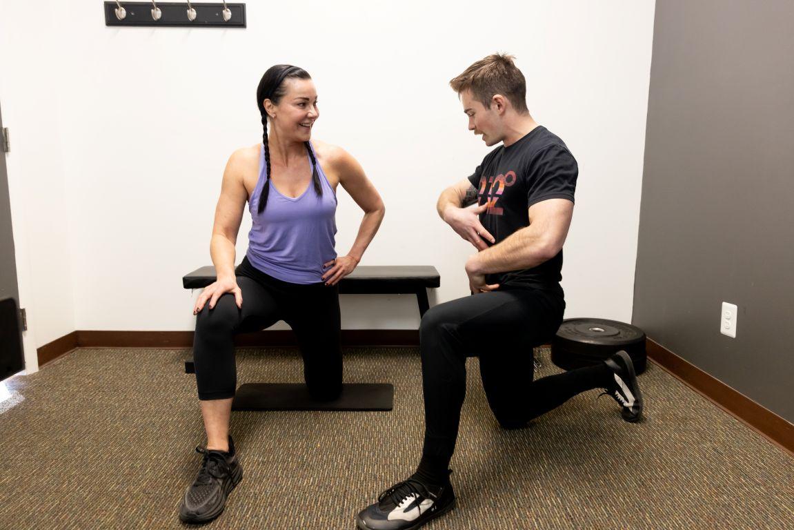 A fitness trainer demonstrates a kneeling lunge exercise to a woman.