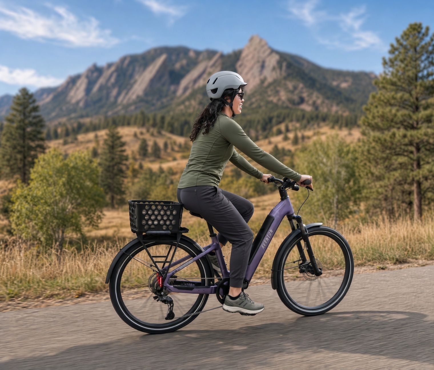 Woman in a helmet riding a purple e-bike with a basket on a paved path, with mountains and trees in the background.