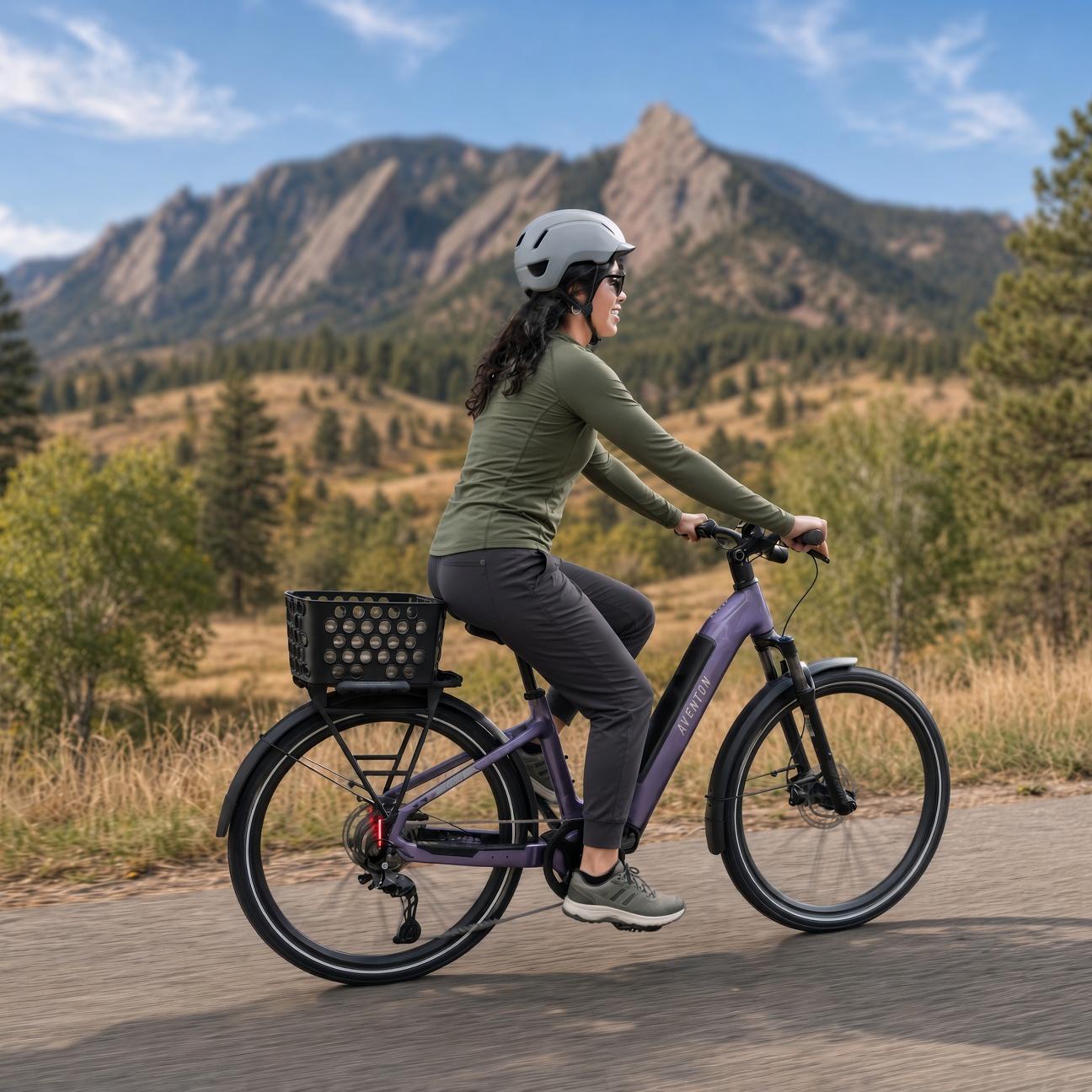 Woman in a helmet riding a purple e-bike with a basket on a paved path, with mountains and trees in the background.